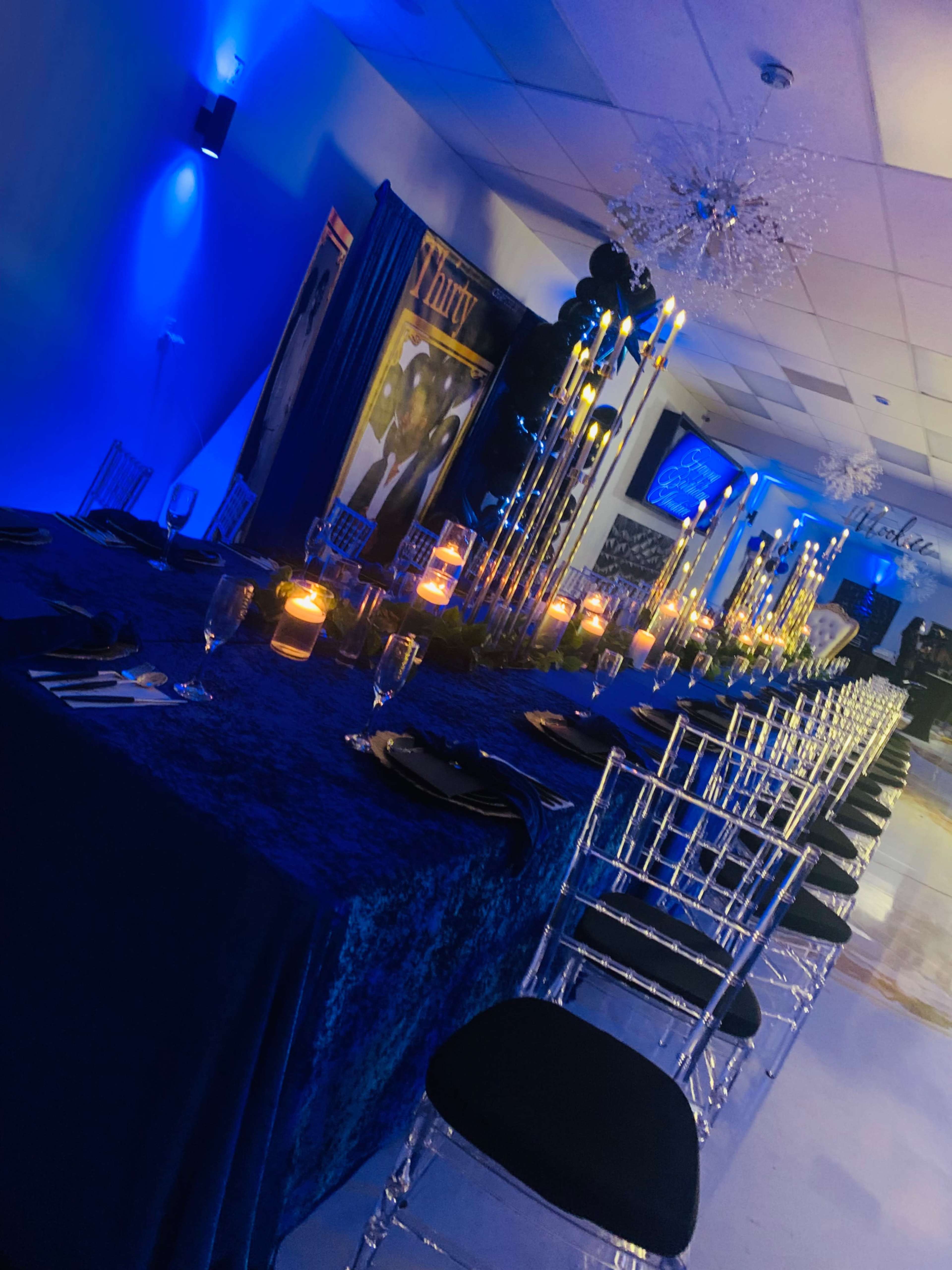 A long dining table is set with candles and elegant black chairs in a dimly lit room with blue lighting and decorative elements.