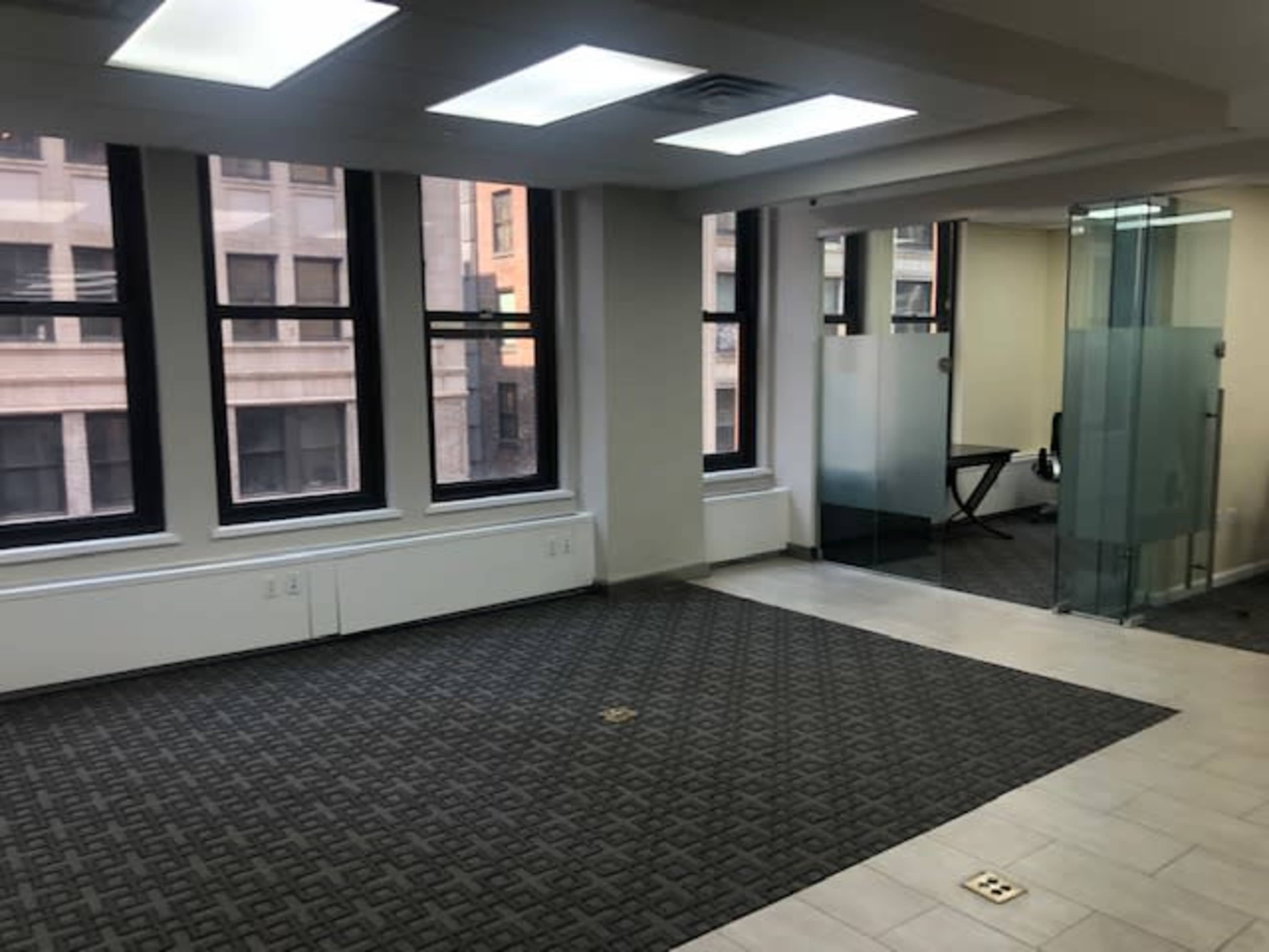 The image shows an empty office space with large windows, gray carpet, and an adjacent glass-walled conference room.