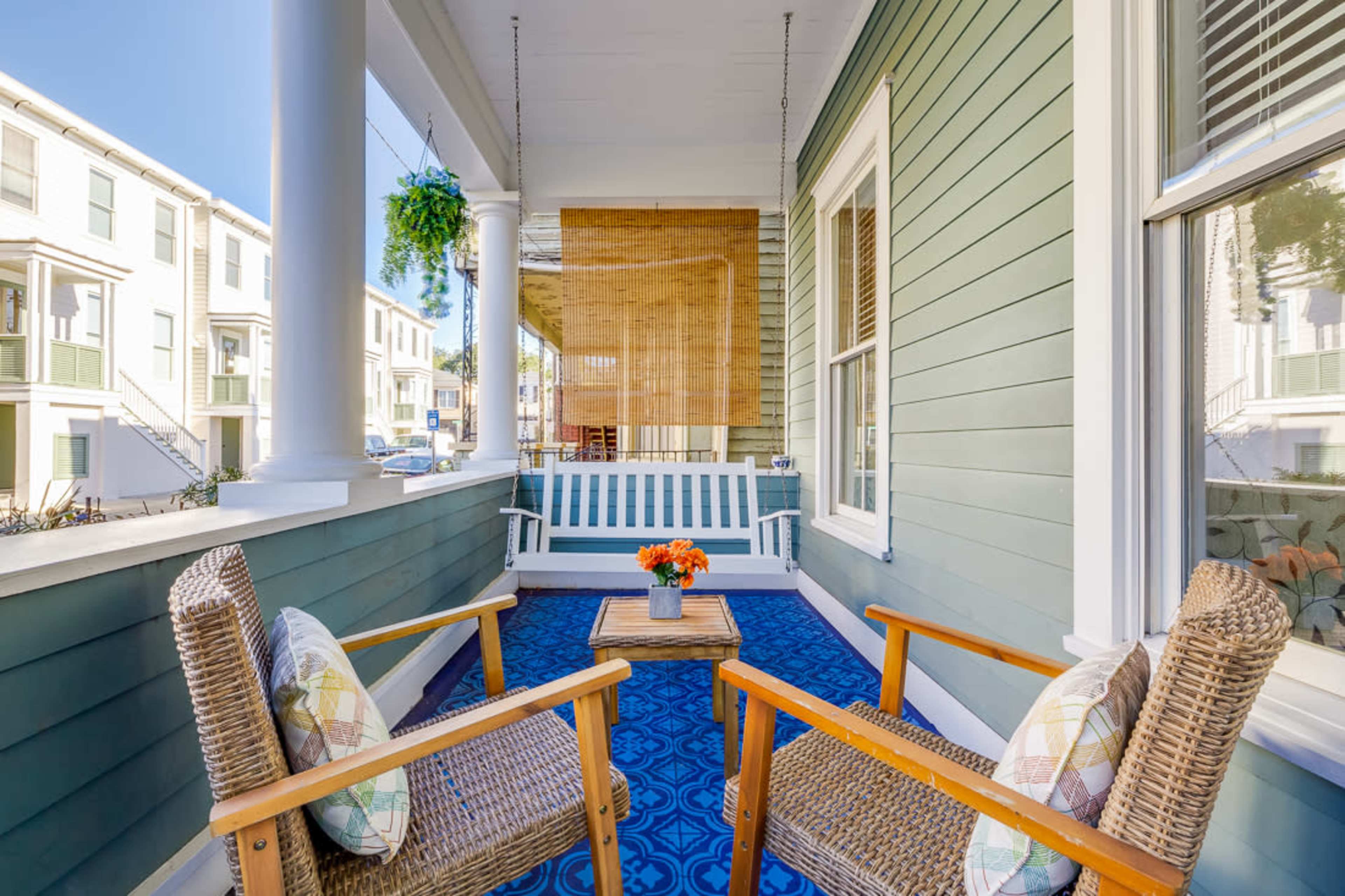 A porch with two wooden chairs, a small table with flowers, and a blue rug, surrounded by green wooden walls and a sunny exterior.