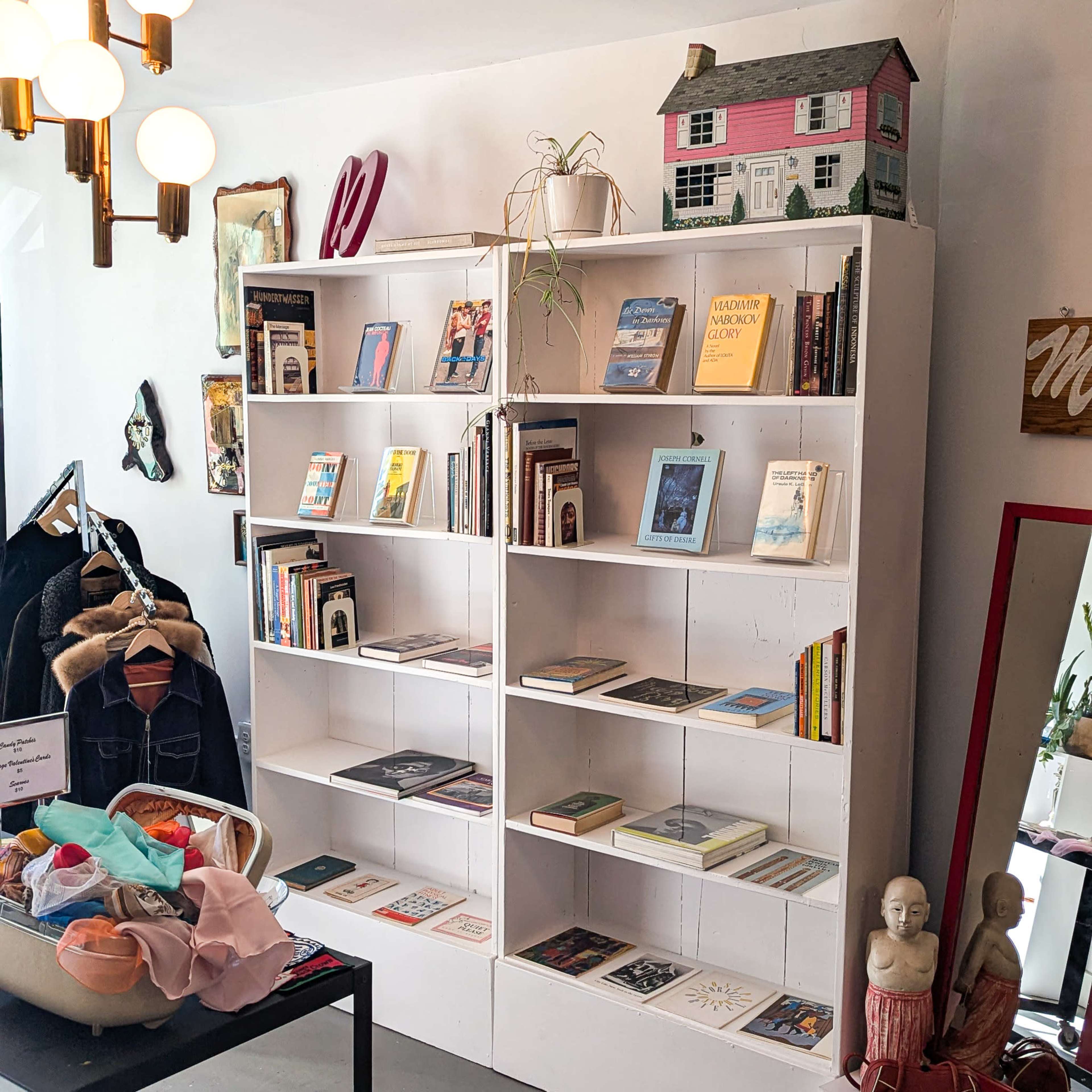 The image shows a white bookshelf filled with books and decorative items, accompanied by a clothing rack and a large mirror in a brightly lit room.