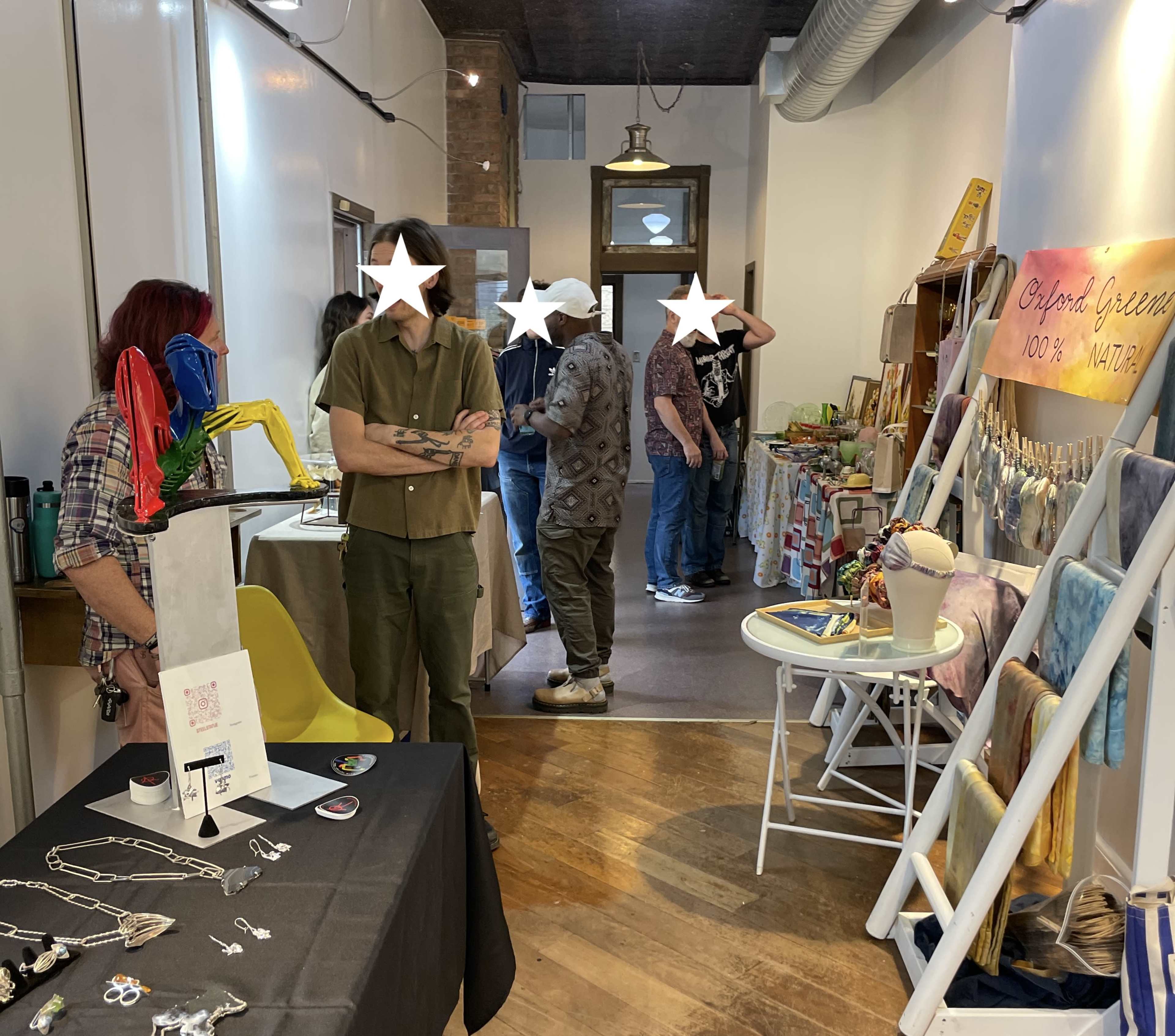 The image shows an indoor market scene with vendors displaying handmade jewelry and crafts, and a few visitors exploring the offerings.