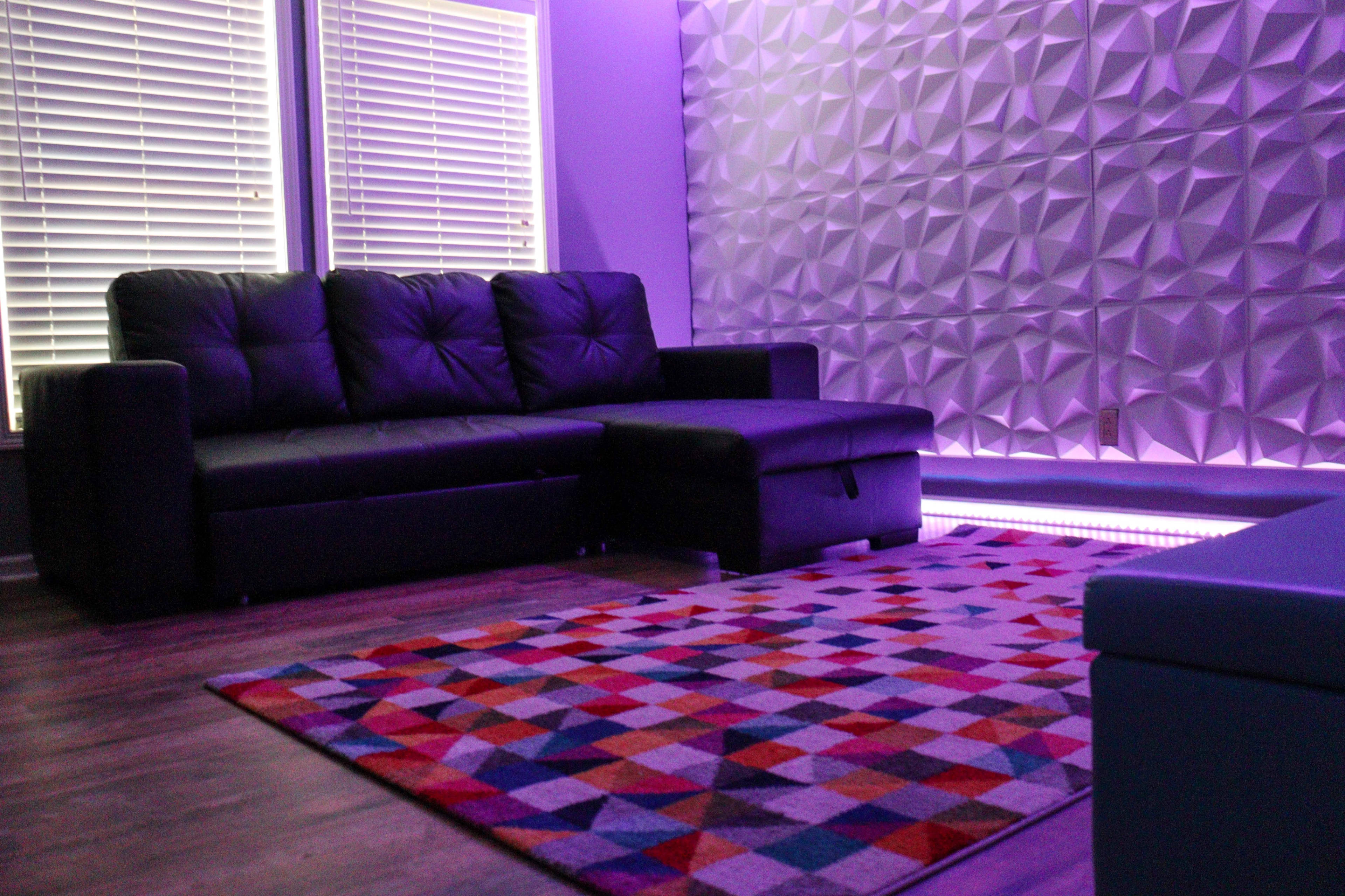 A modern living room featuring a black sectional sofa, a patterned area rug, and a wall with a geometric design illuminated by purple lighting.