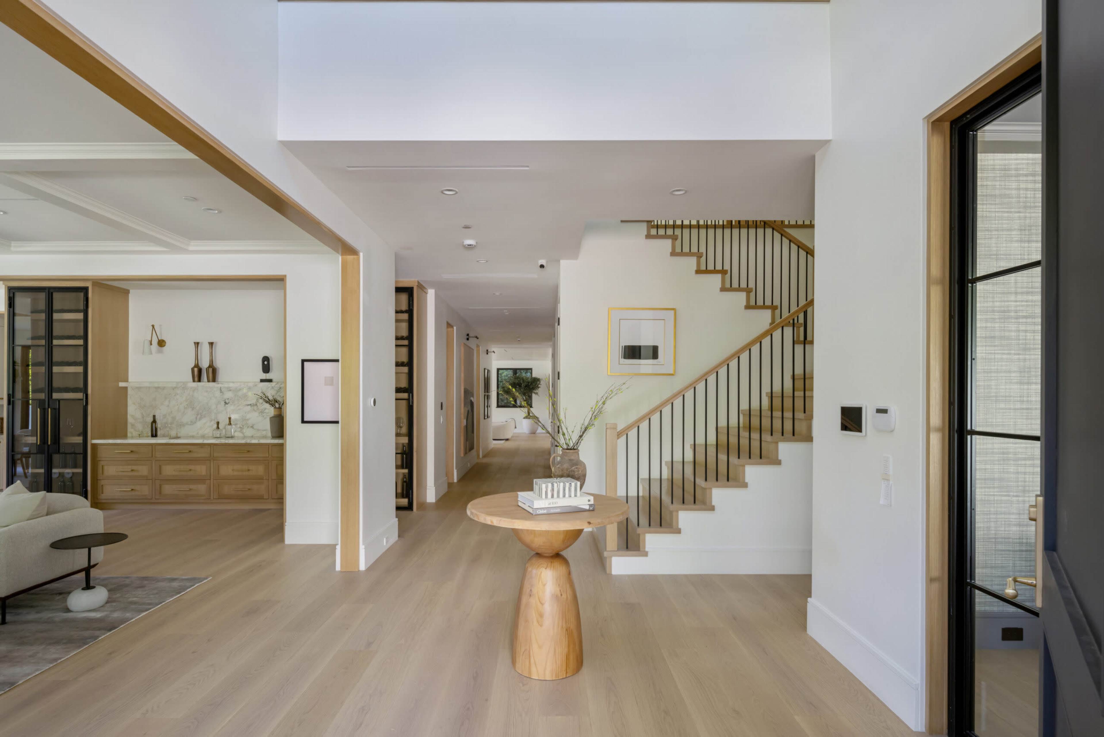 The image shows a modern entryway with a circular wooden table, a staircase with a railing, and minimalist decor featuring light-colored walls and flooring.