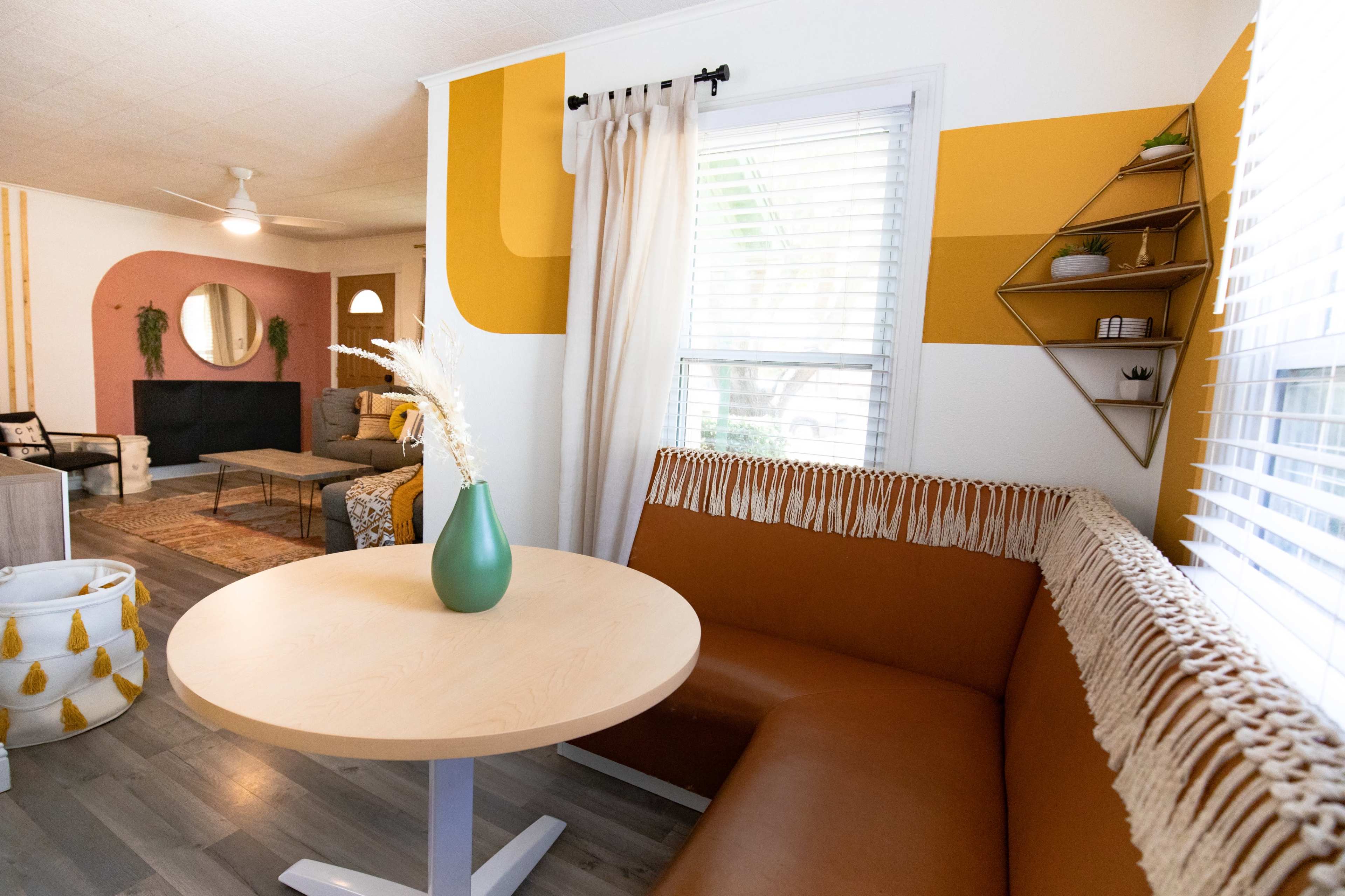 A cozy dining area with a round table and a curved, leatherette bench featuring fringe, set against a wall painted with geometric patterns in warm colors.