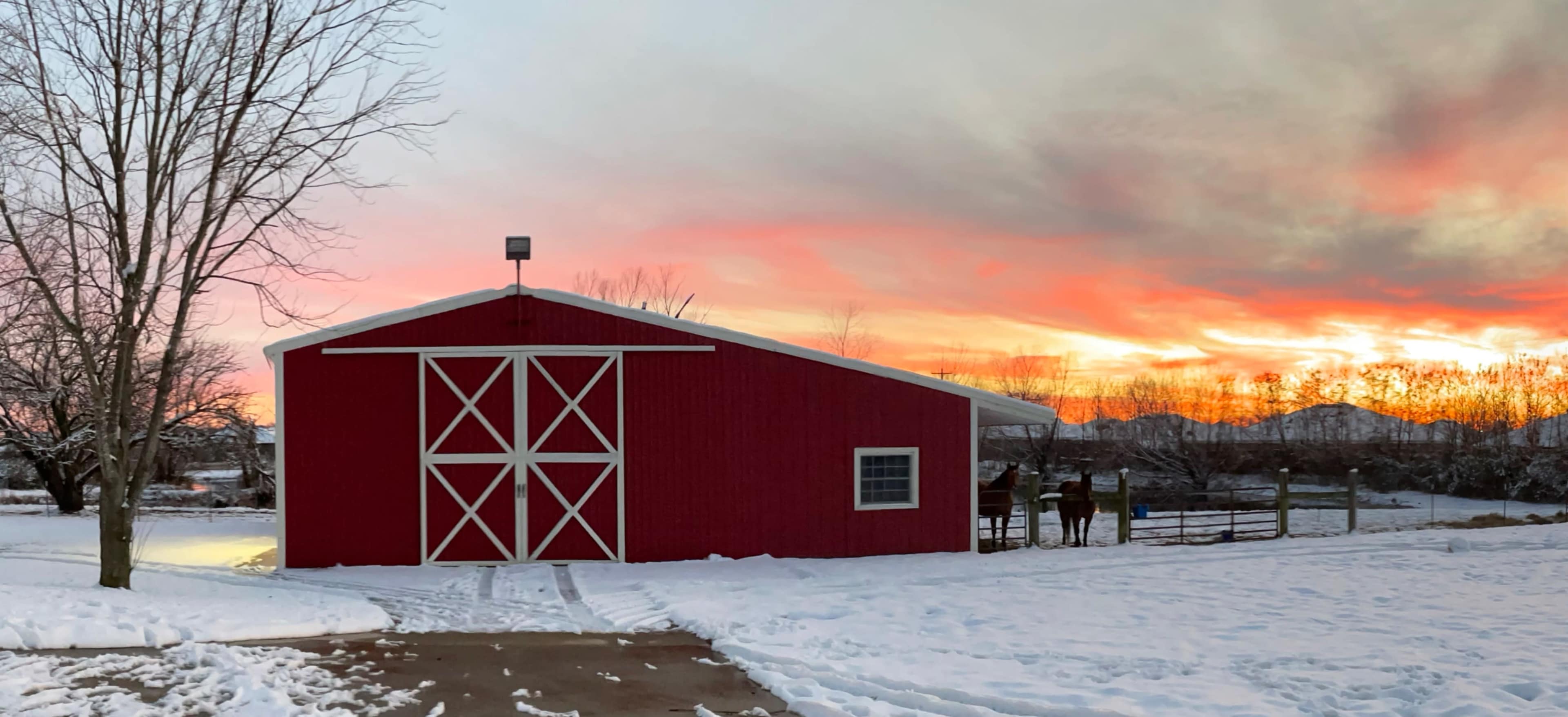A red barn stands amidst a snowy landscape as a vibrant sunset illuminates the sky behind it.
