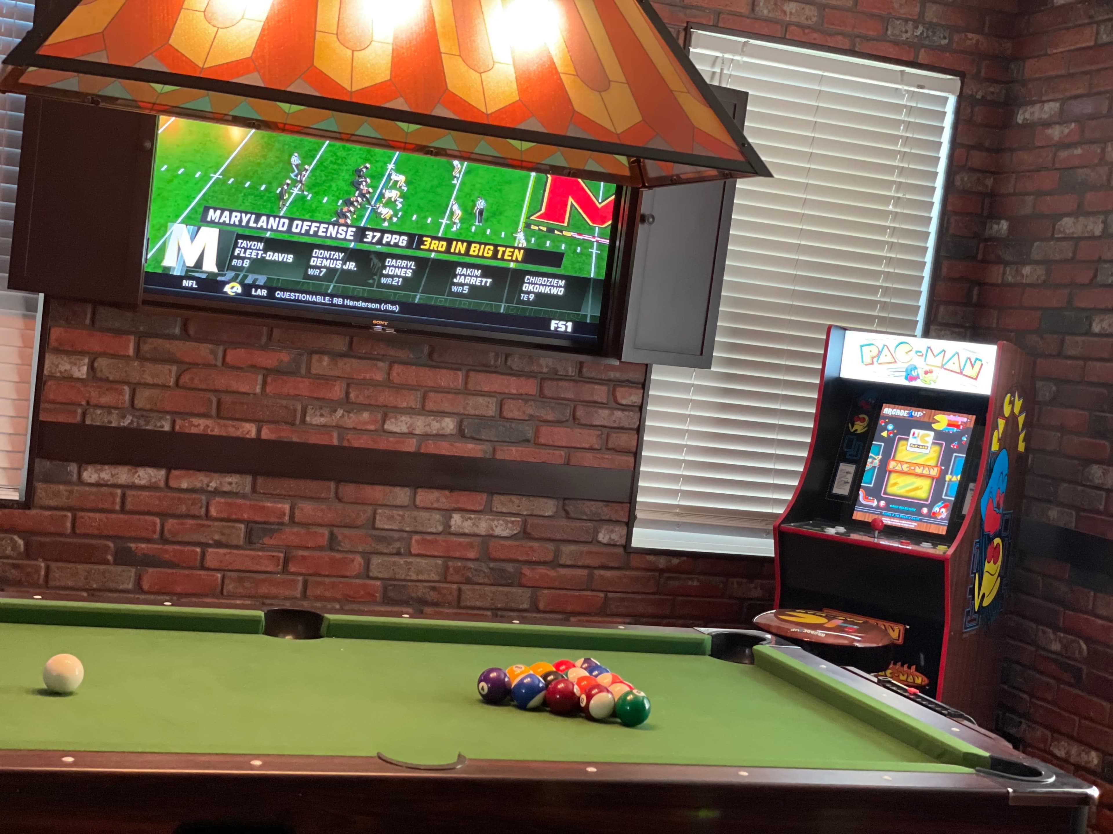 A green pool table with a triangle of balls sits under a patterned lampshade, while a vintage Pac-Man arcade game and a television displaying a football game are nearby.