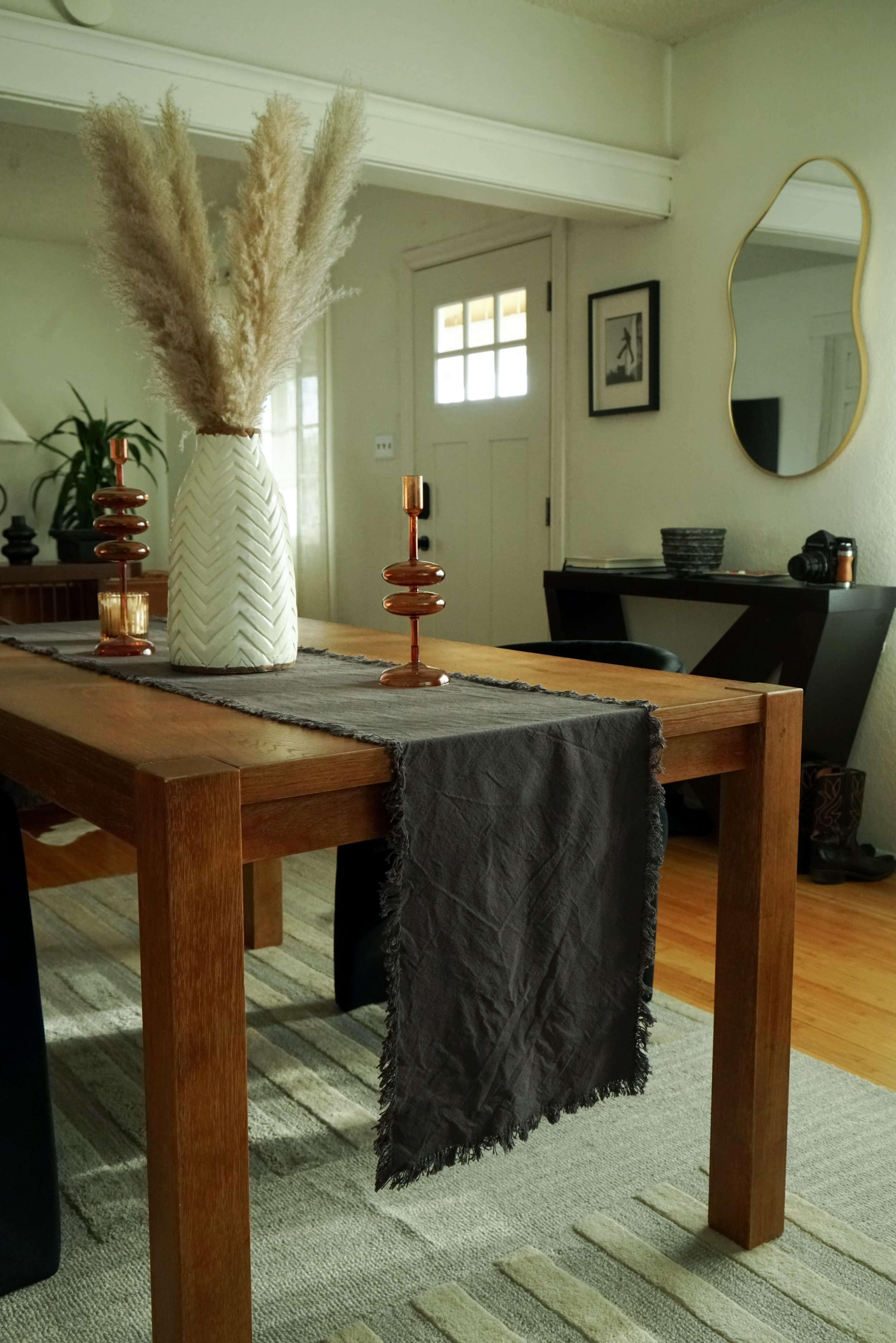 A wooden dining table is centrally positioned in a well-lit room, adorned with a dark table runner, a textured vase filled with pampas grass, and decorative candles.