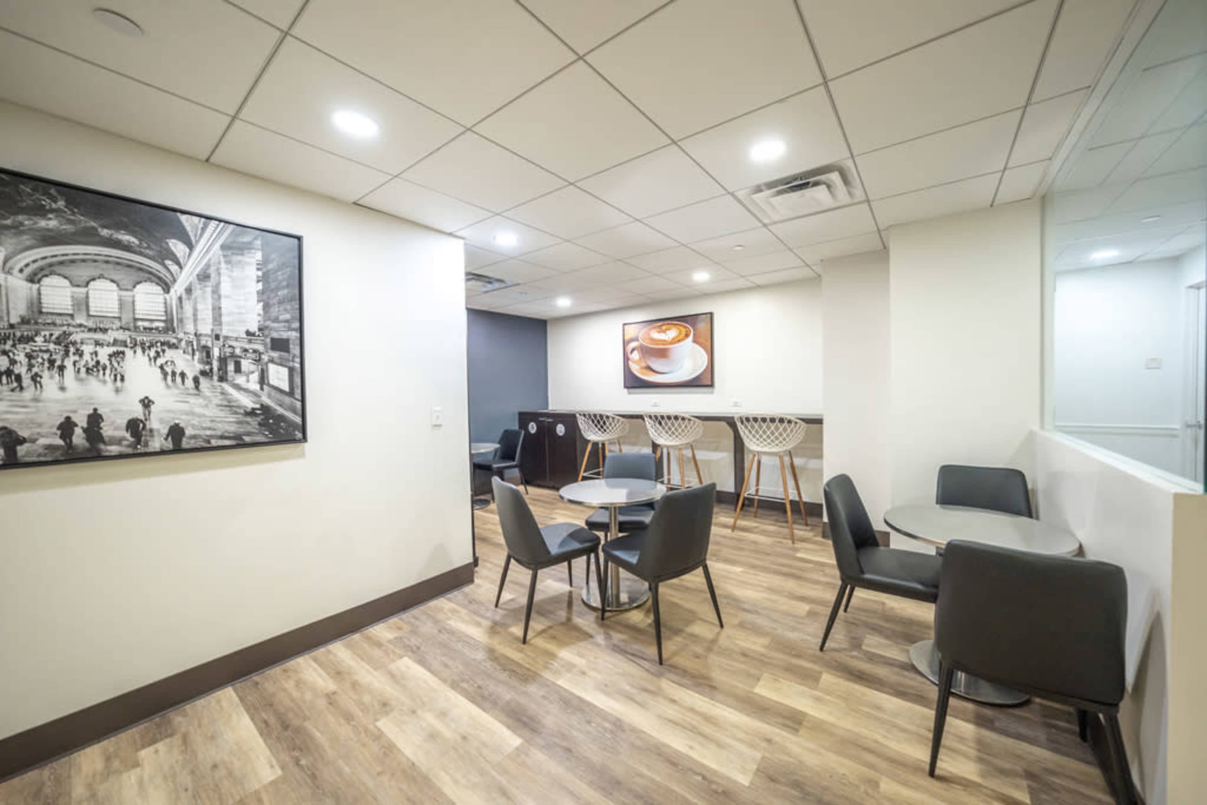The image shows a modern, minimalistic lounge area with tables and chairs, featuring two wall-mounted art pieces.