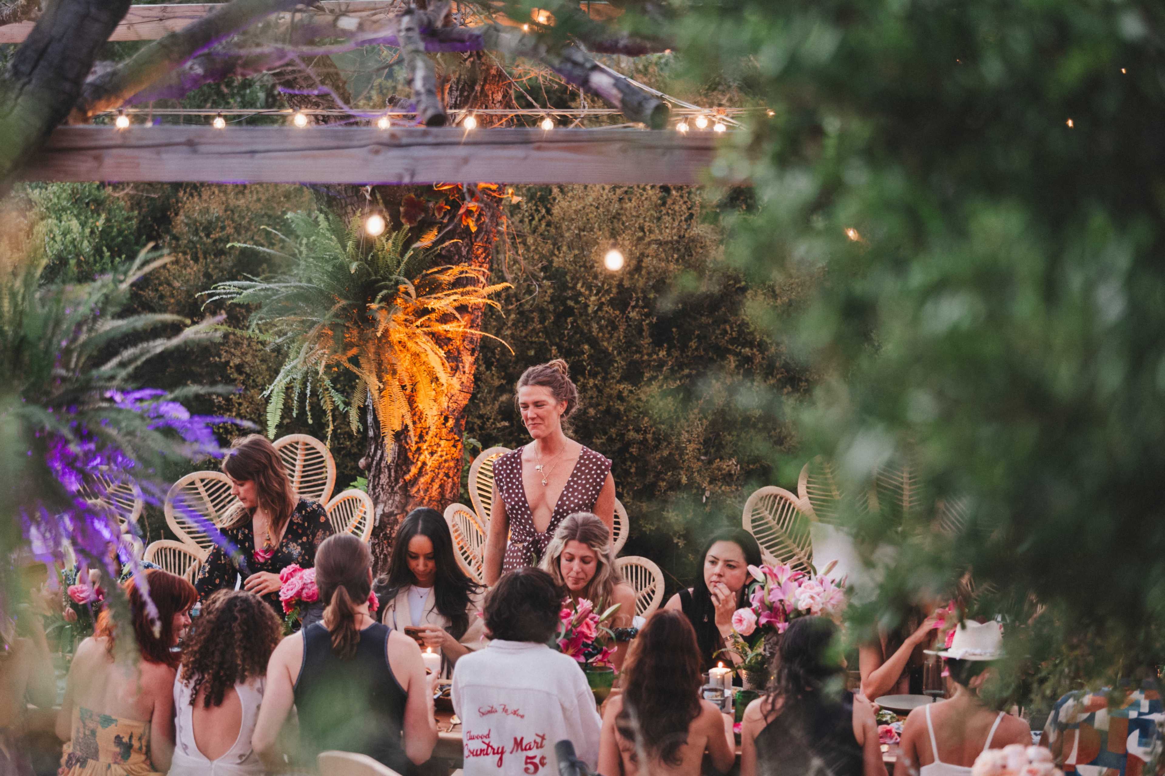 A group of people gathered around a decorated table in an outdoor setting with plants and string lights.