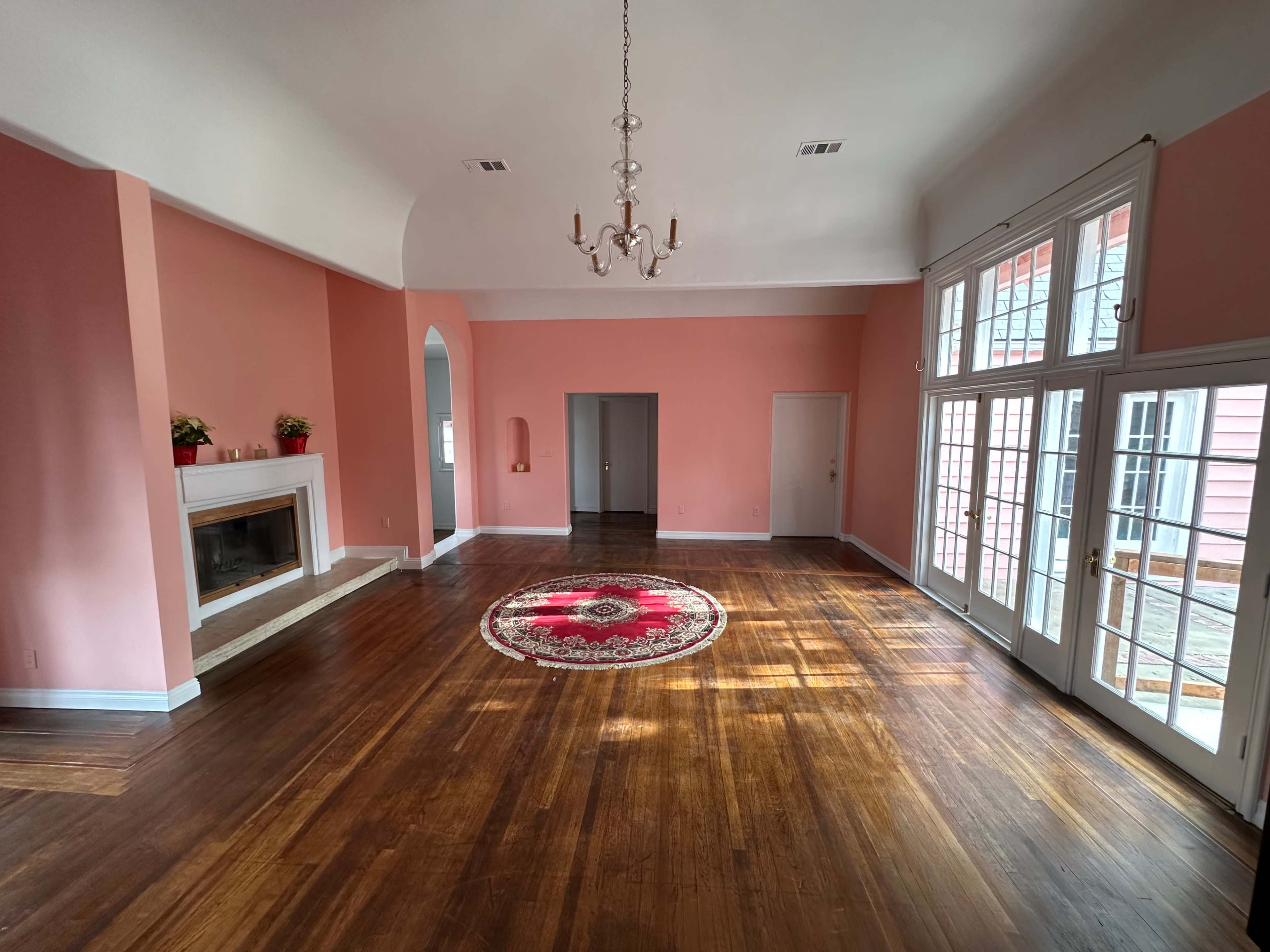 The room features a pink wall design, a circular area rug in the center, hardwood floors, a fireplace, and large windows with French doors leading to an outdoor space.