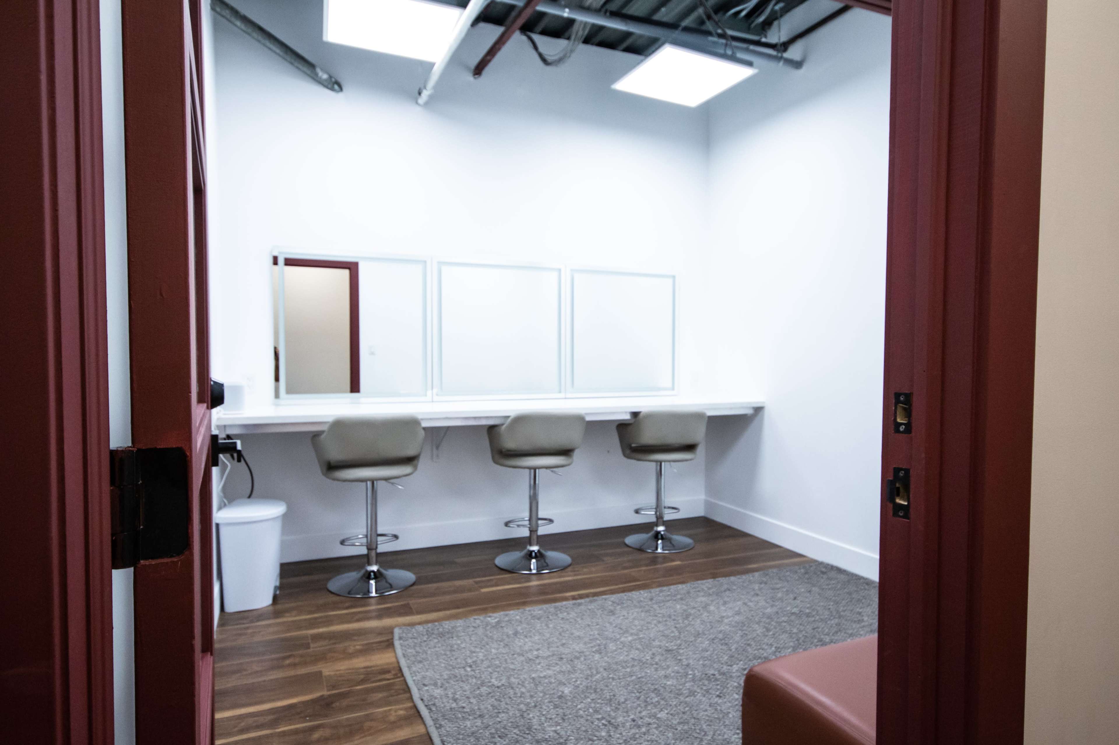 A brightly lit dressing room features three bar stools in front of a long mirror and a small waste bin in the corner.