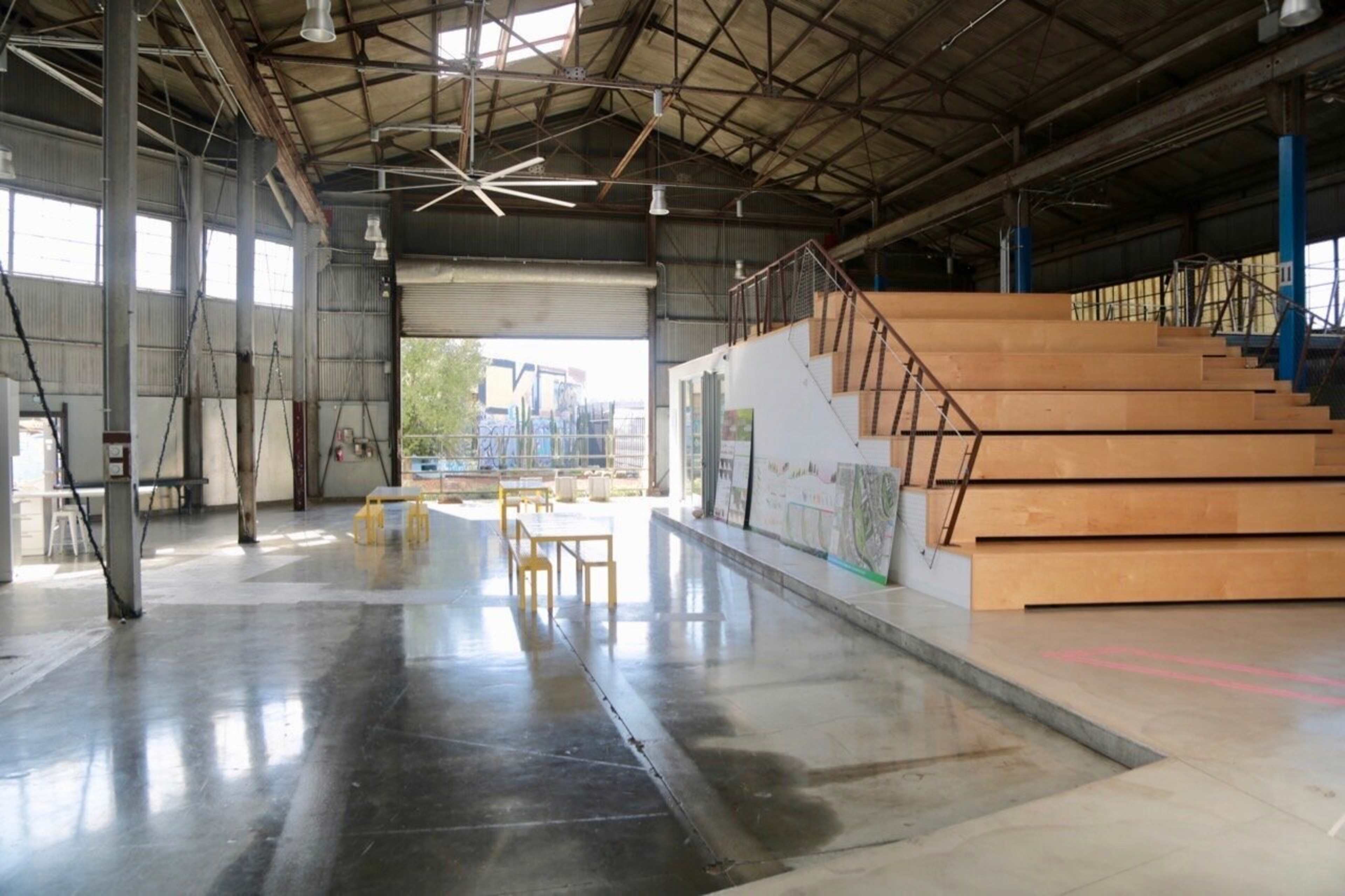 The image shows an industrial-style interior with wooden steps leading to a platform, surrounded by large windows and minimalist furniture.