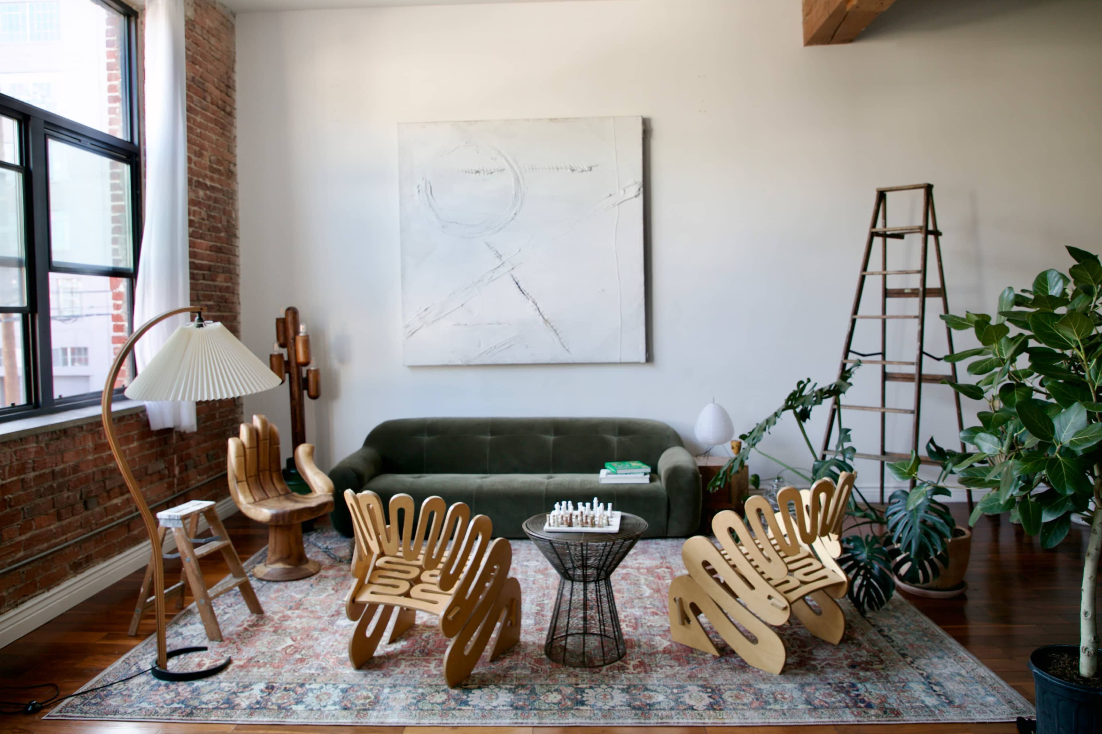 A modern living room featuring a green sofa, unique wooden chairs, a small table with a chess set, and various decorative plants.