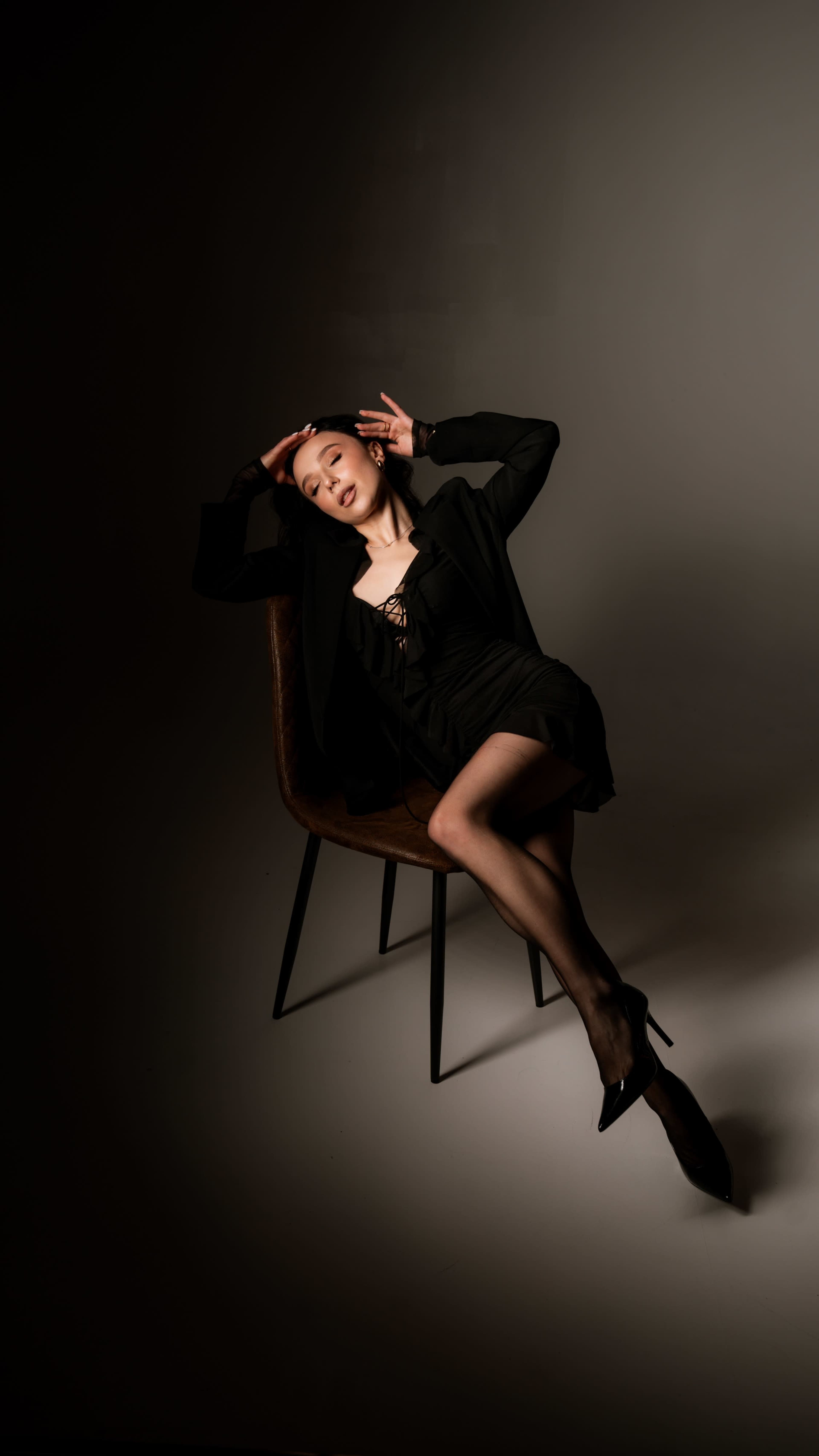 A person in a black outfit poses casually while seated on a chair, with one hand touching their head and the other resting on their lap.