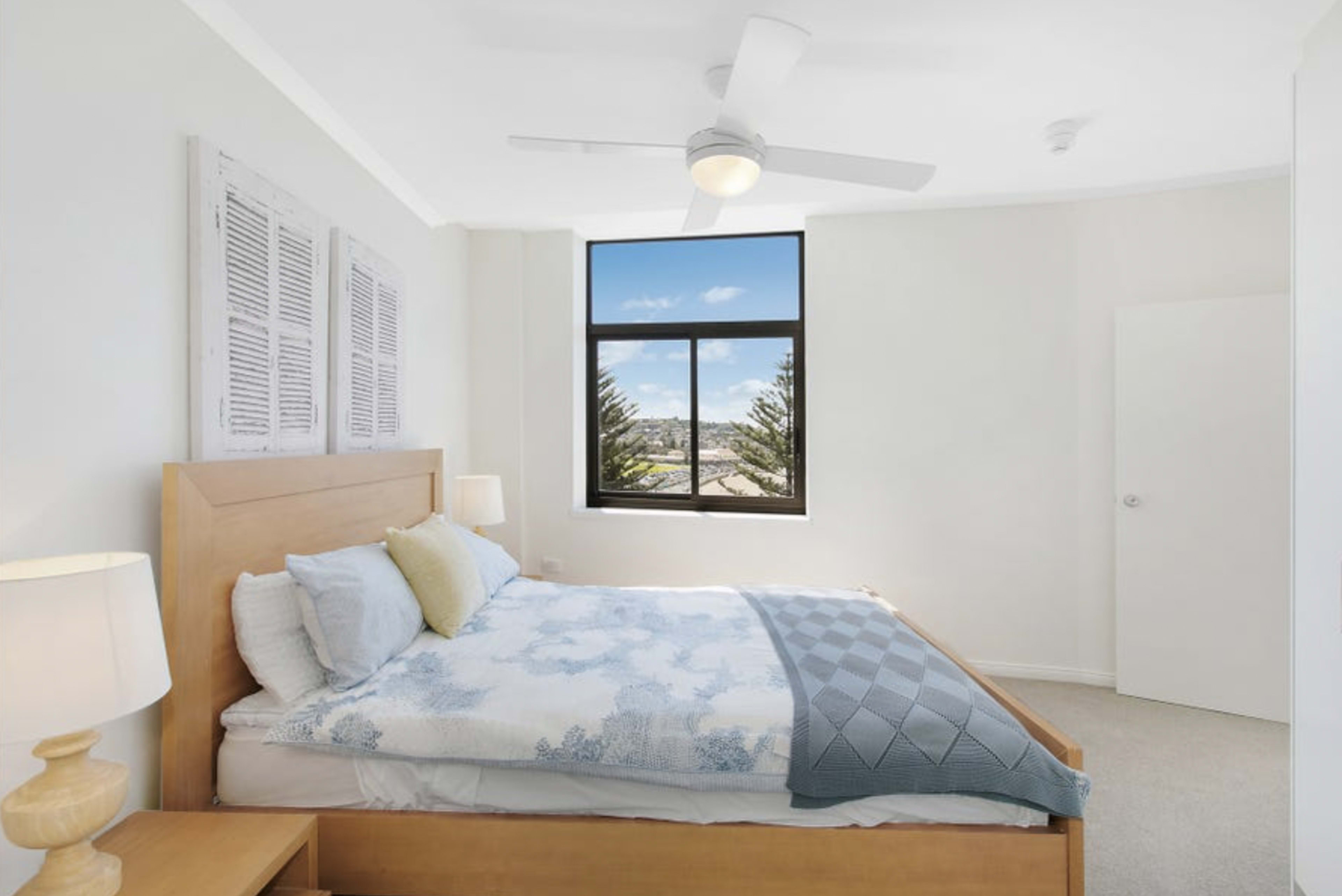A bright bedroom with a wooden bed, light-colored bedding, a ceiling fan, and a large window overlooking a view of trees.