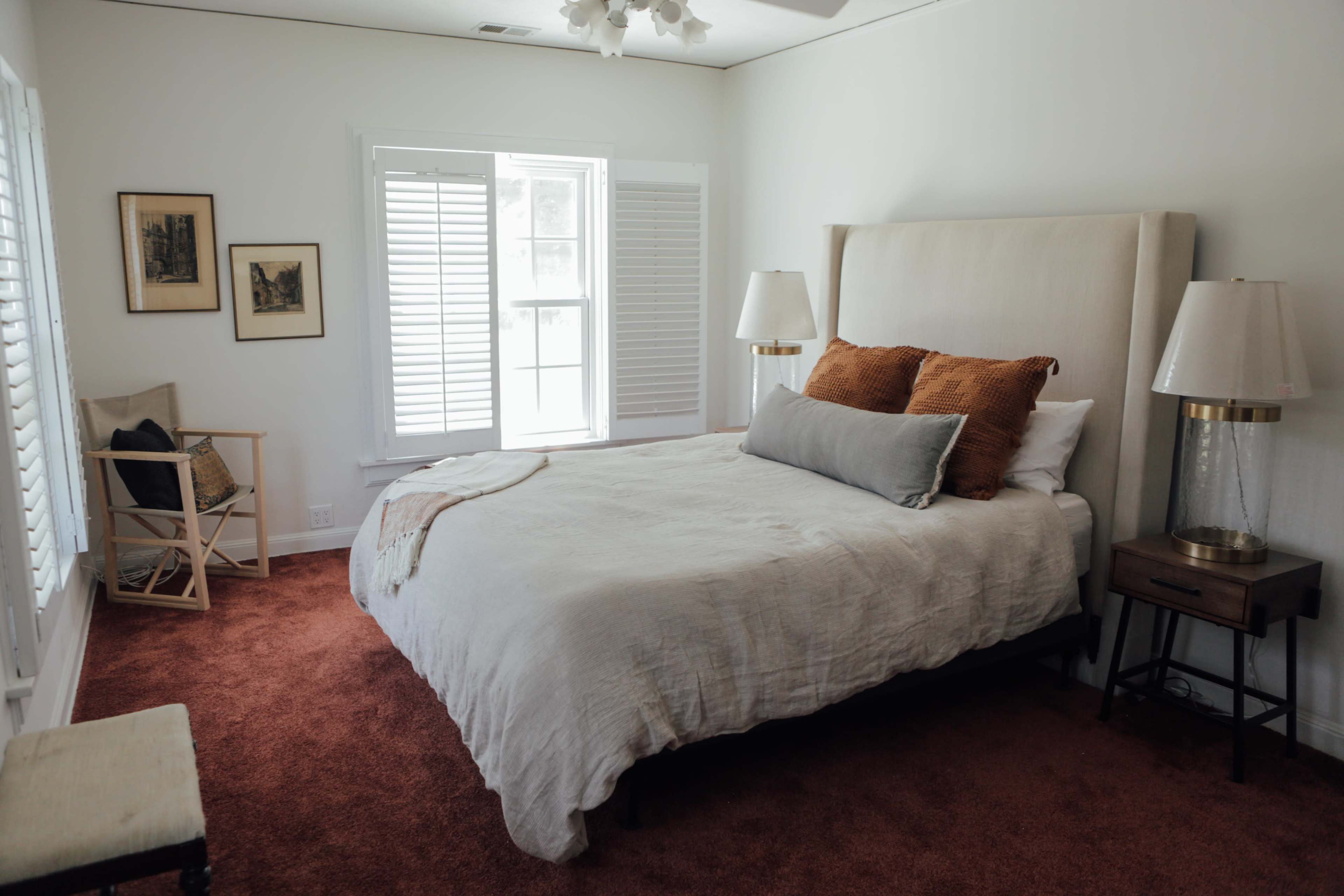 The image shows a bedroom with a bed dressed in neutral linens, two bedside lamps, and a window with white shutters.