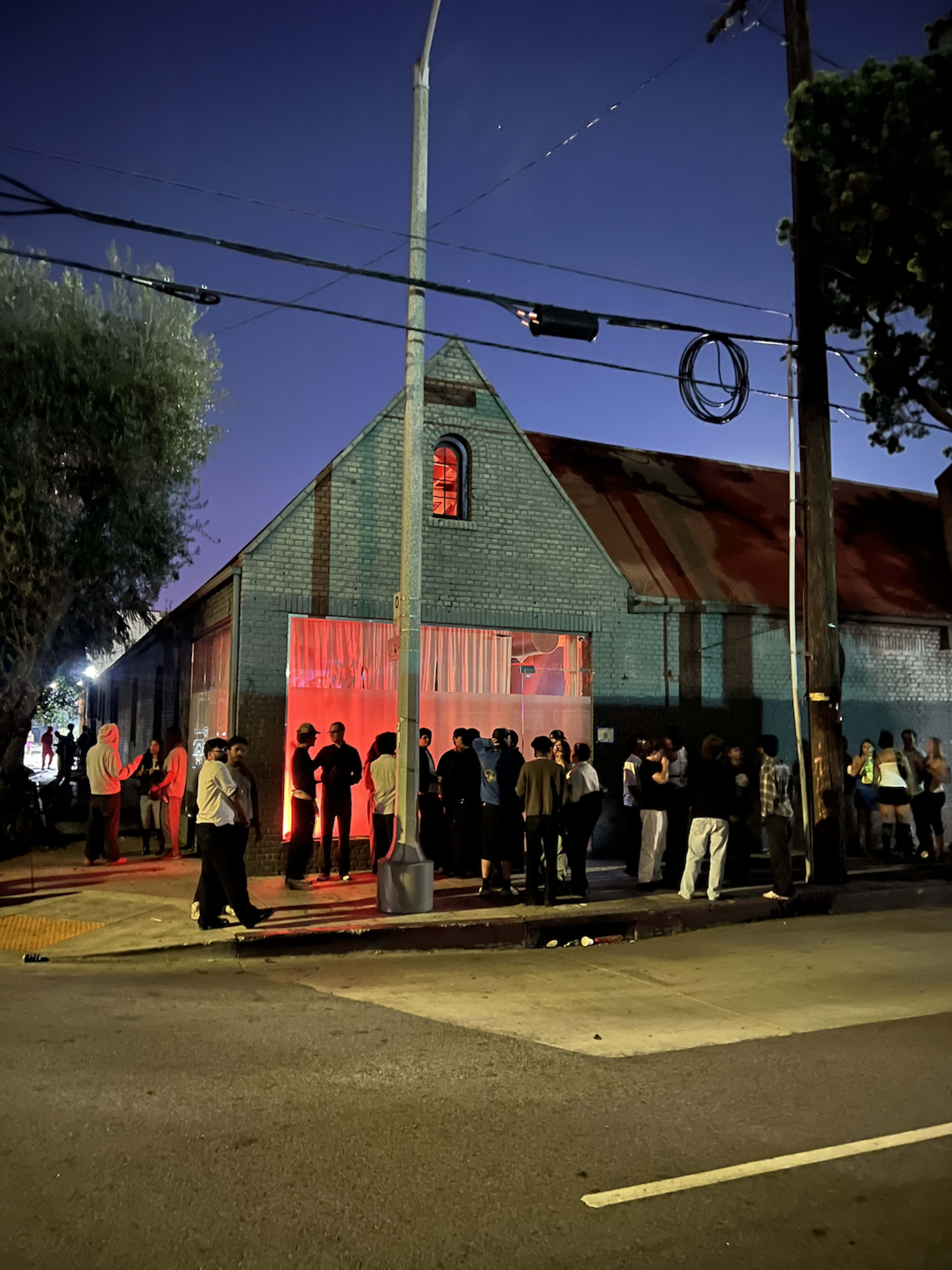 A crowd of people gathers outside a converted warehouse with a lit entrance at dusk.