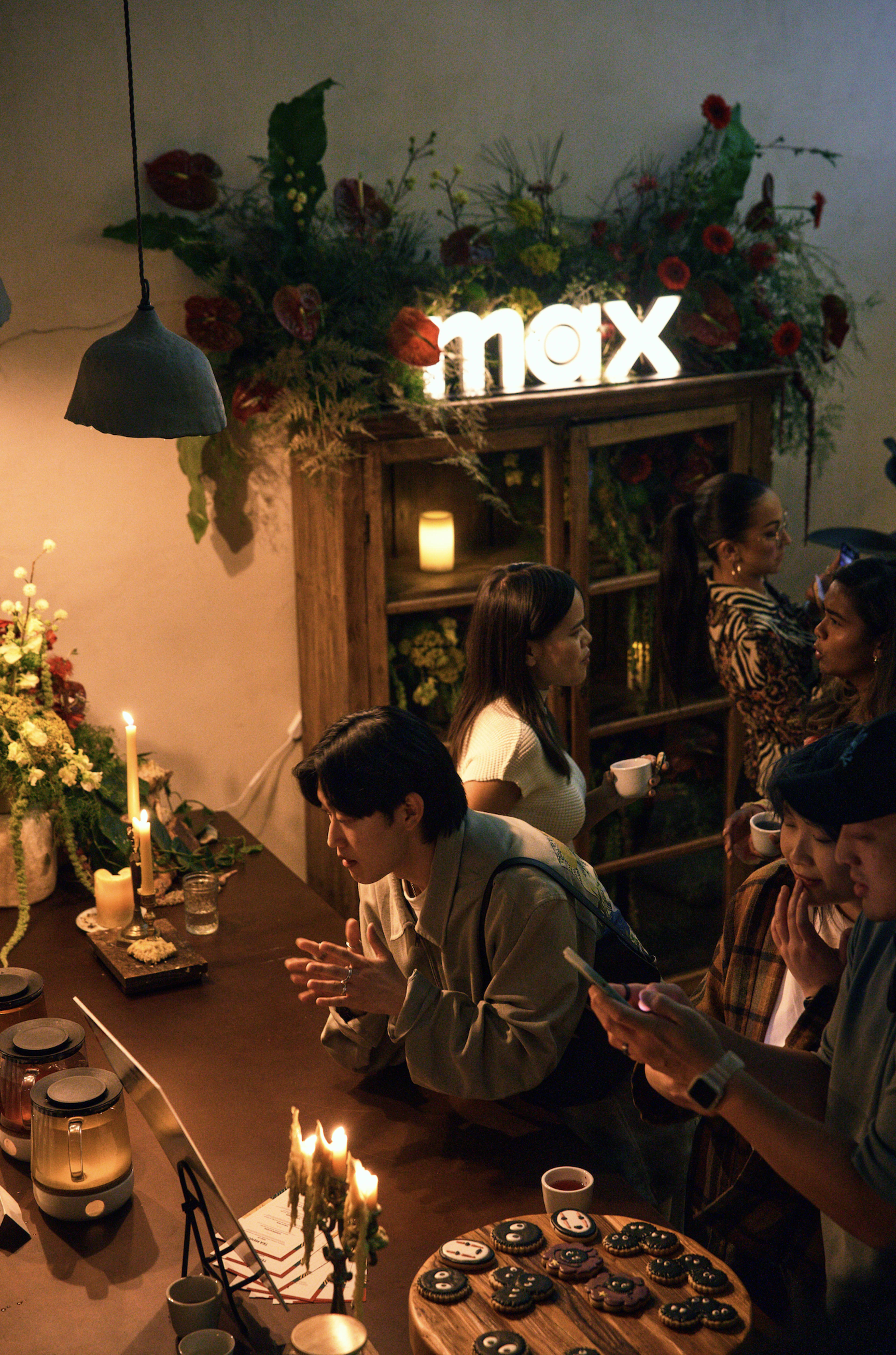 A group of people is gathered around a table in a cozy, flower-decorated café, engaging in conversation and looking at a laptop.