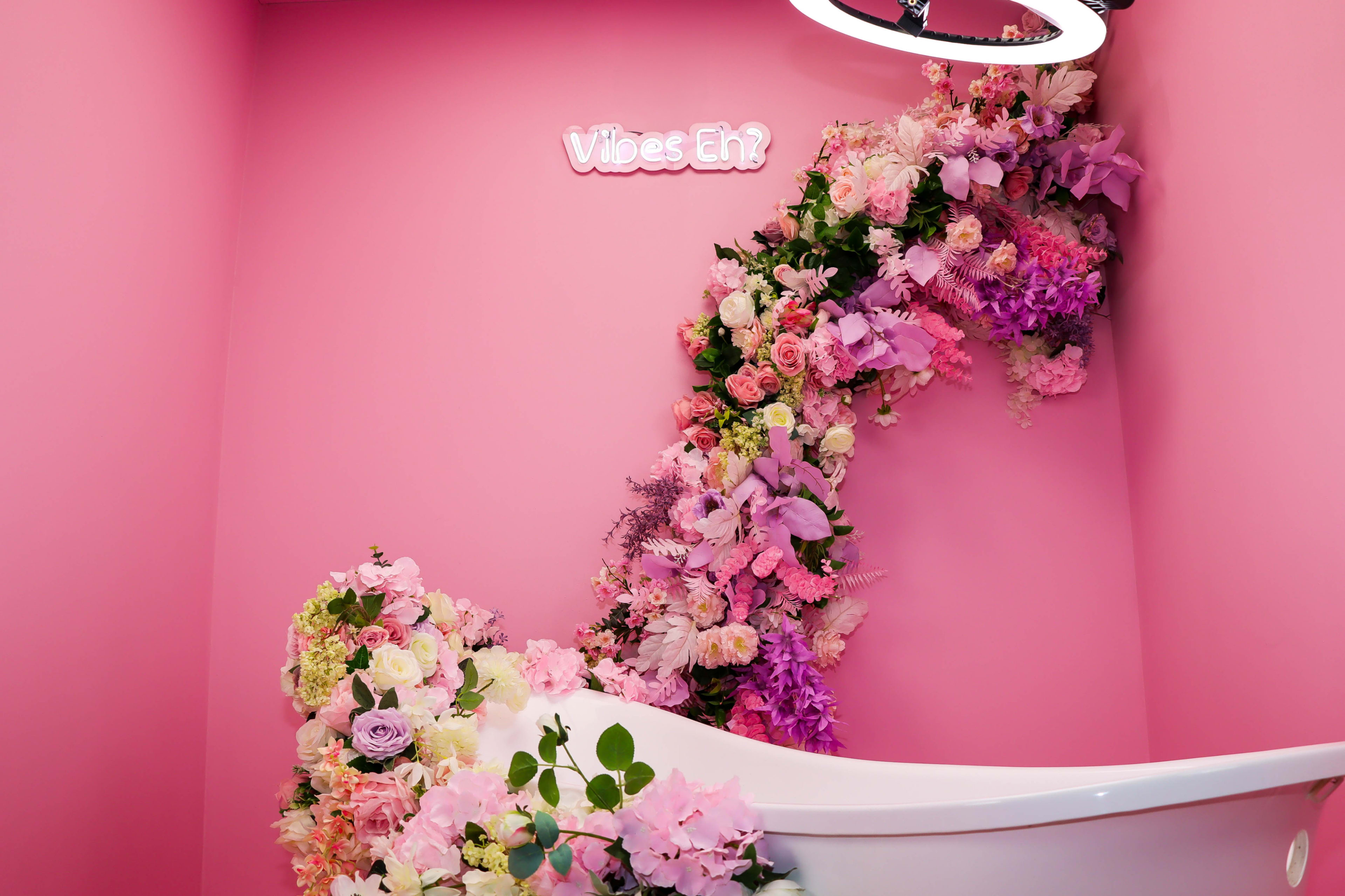 A white bathtub is positioned against a pink wall adorned with a colorful floral arrangement that curves towards a sign reading "Vibes Eh?"