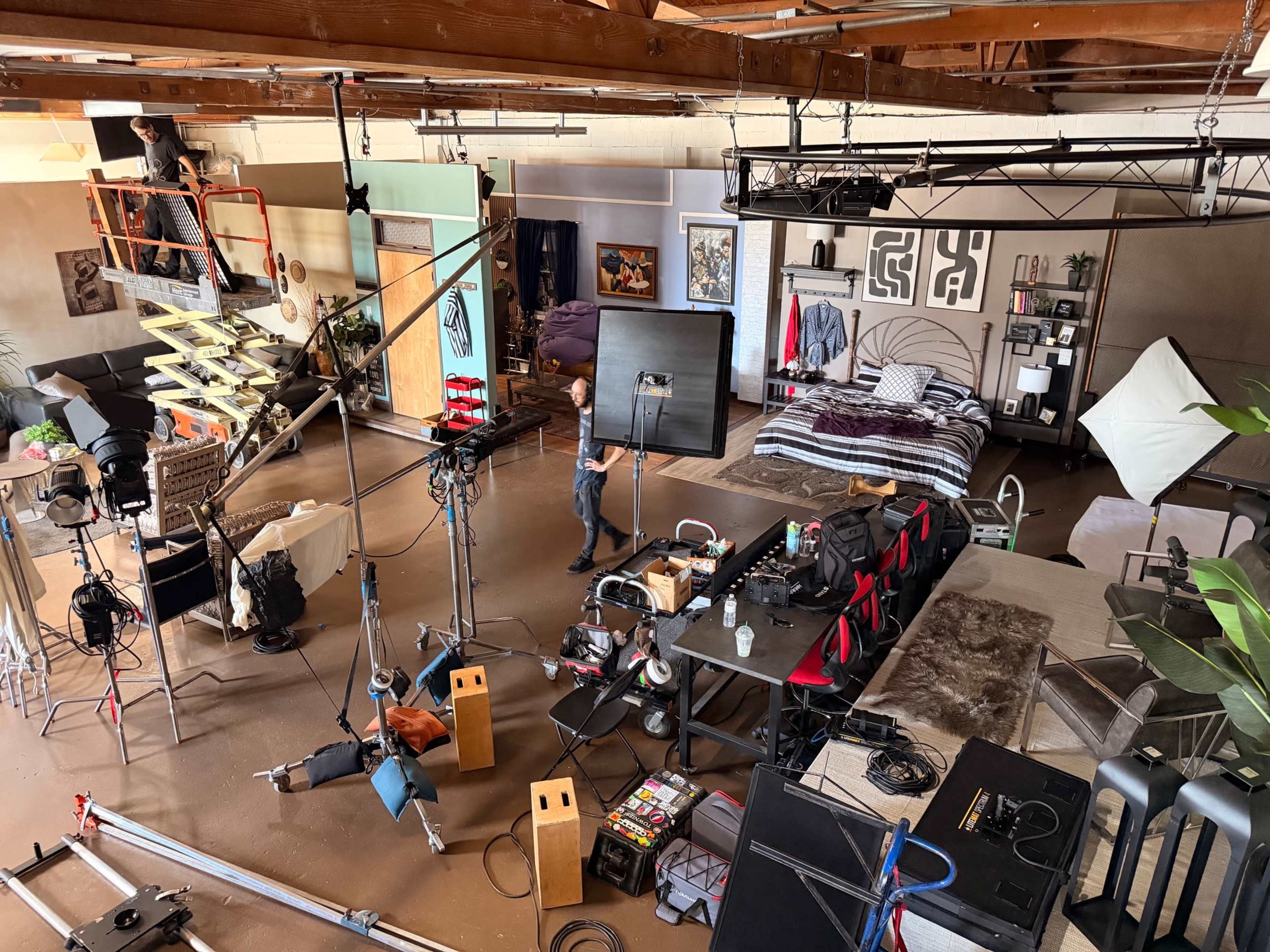 The image depicts a film or photography studio setup, featuring various equipment, a bed, lighting rigs, and decorative furniture arranged in an open space.