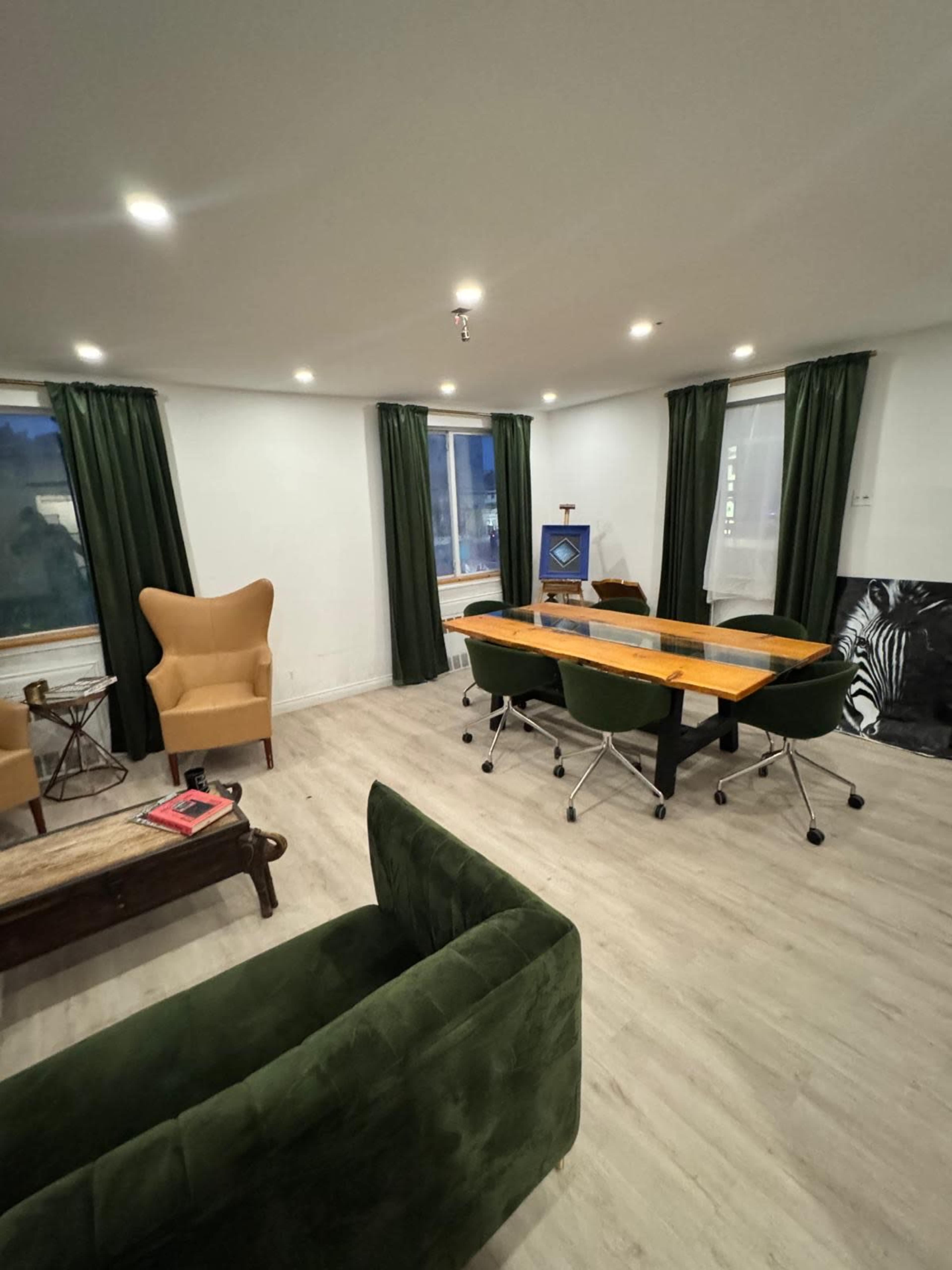 The image shows a modern meeting room featuring a long wooden table surrounded by rolling chairs, a green sofa, and a wingback armchair, with large windows framed by dark green curtains.