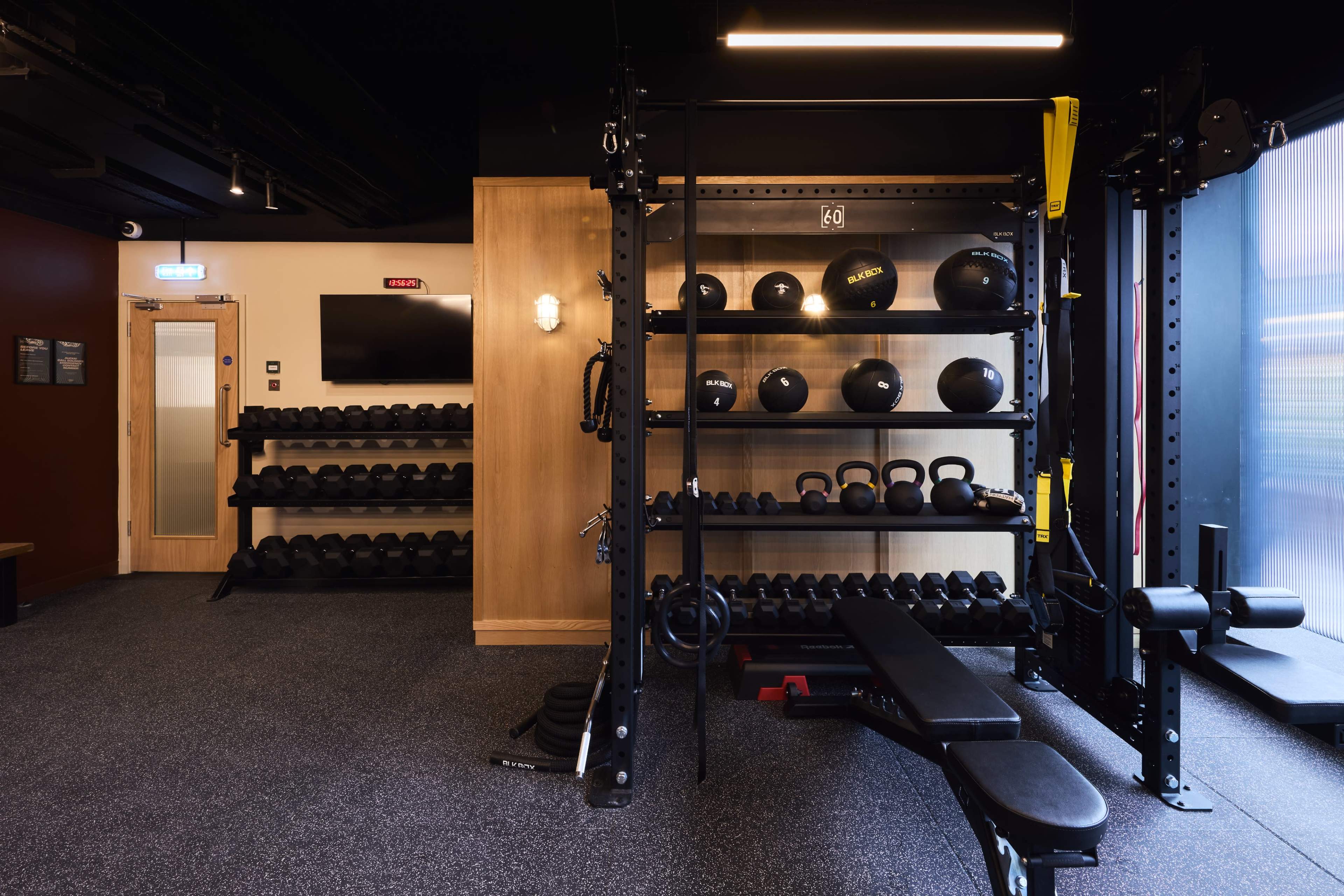 The image shows a well-equipped gym space featuring a shelf of black dumbbells, kettlebells, and a bench press against a modern interior backdrop.