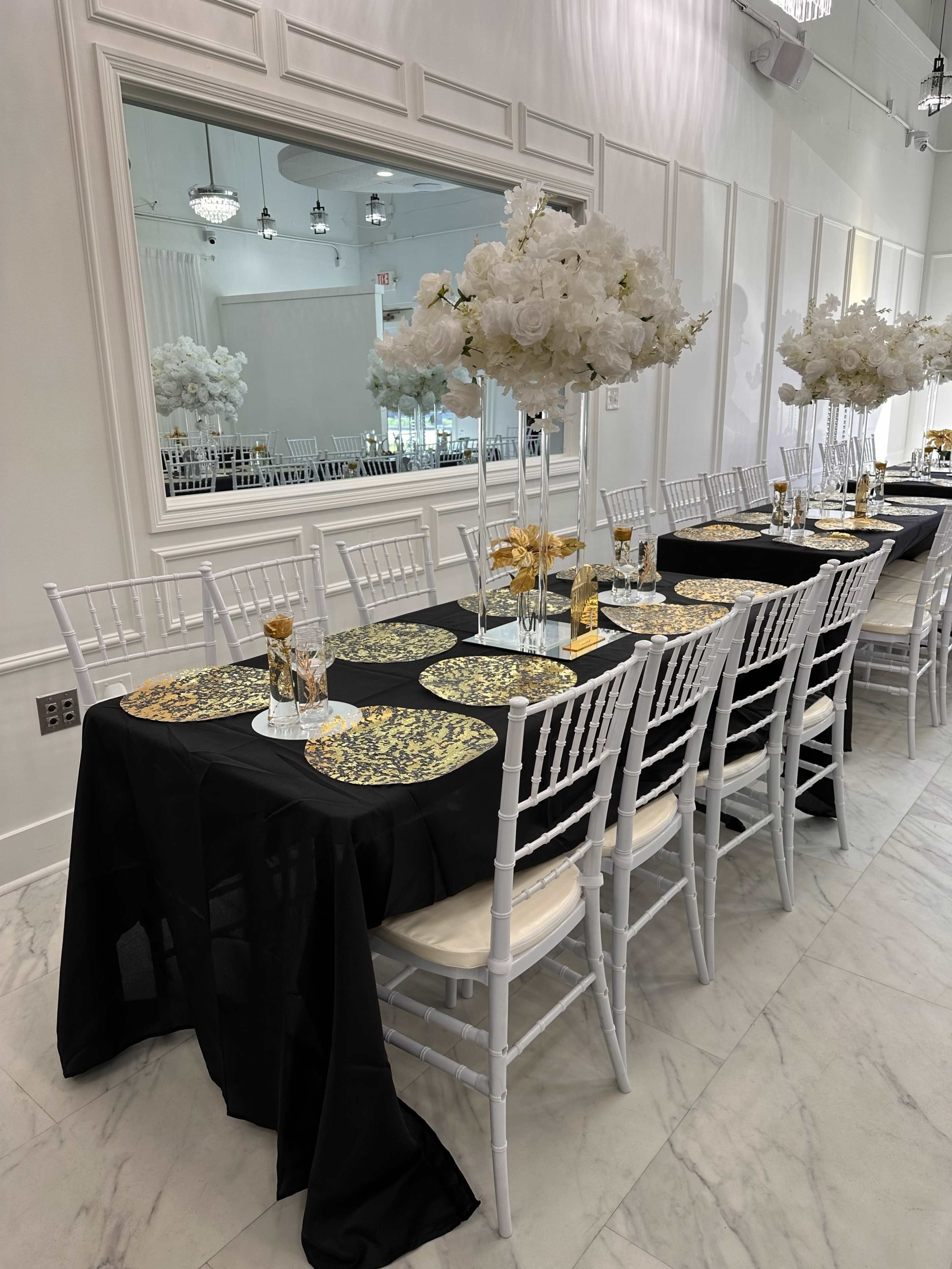 The image shows a formal dining setup with a long black table adorned with gold and white floral centerpieces and elegant place settings.
