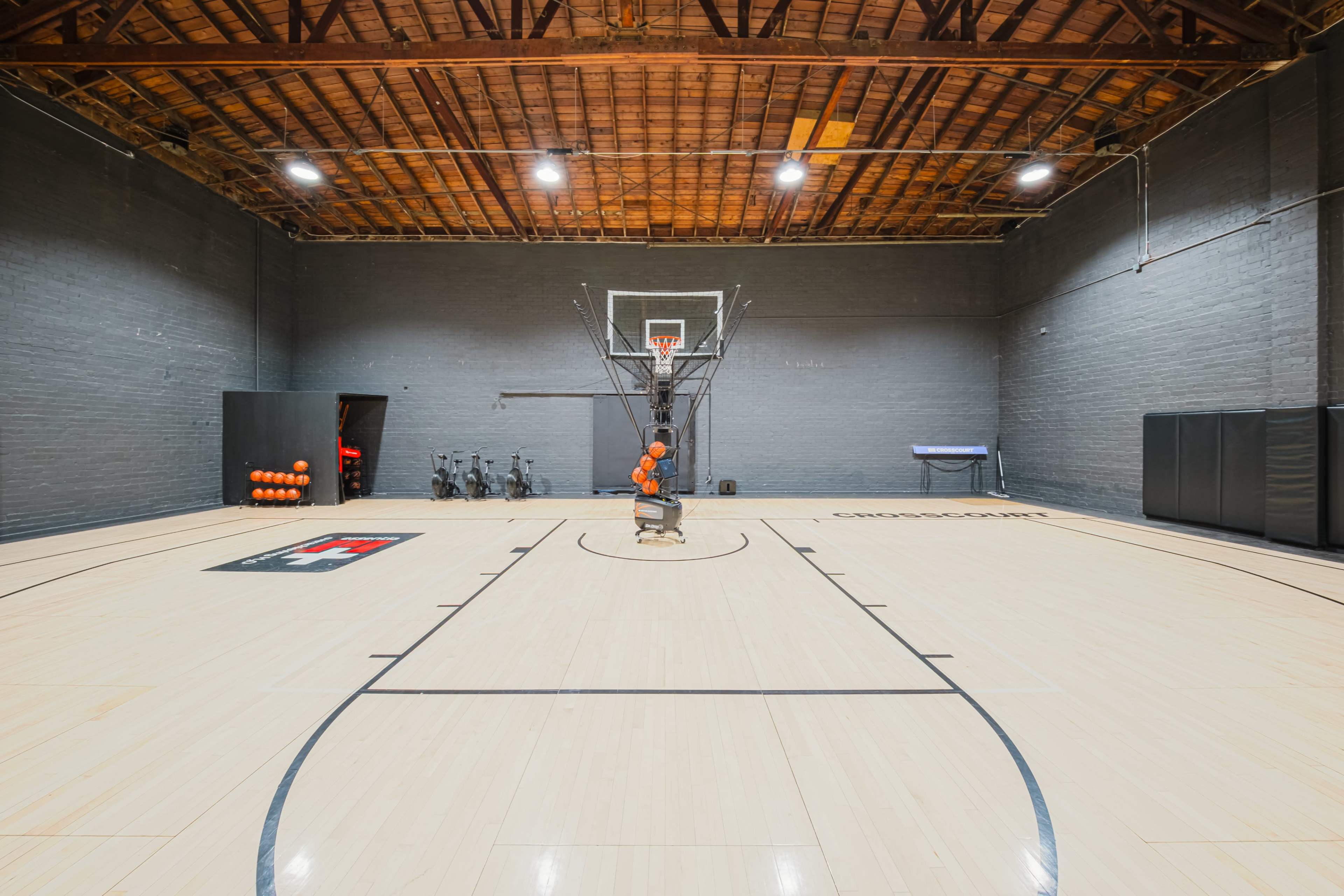 The image shows a basketball court with a hoop, racks of basketballs, and training equipment against a gray wall.