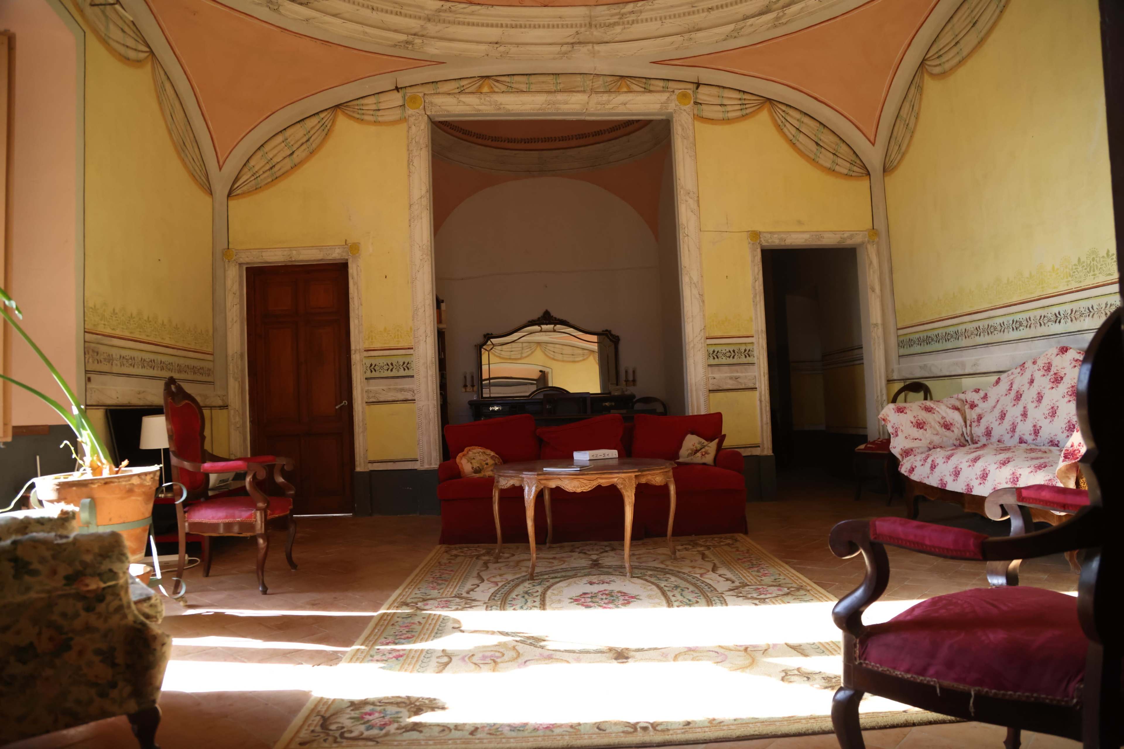 The image shows a spacious room with high ceilings, a central red sofa, wooden doors, and ornate wall decorations.