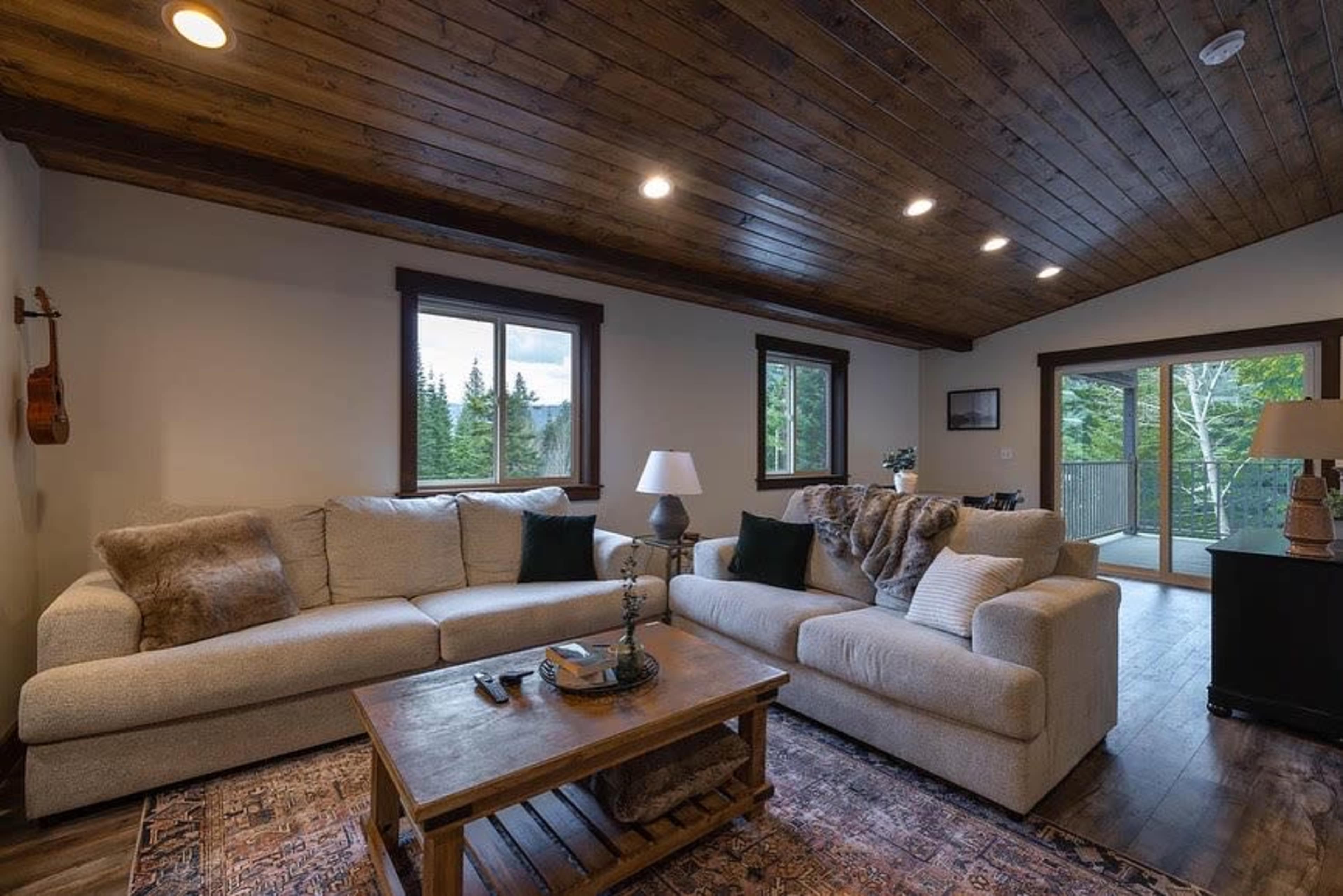 A cozy living room featuring a light-colored sectional sofa, a wooden coffee table, and large windows overlooking a forested landscape.