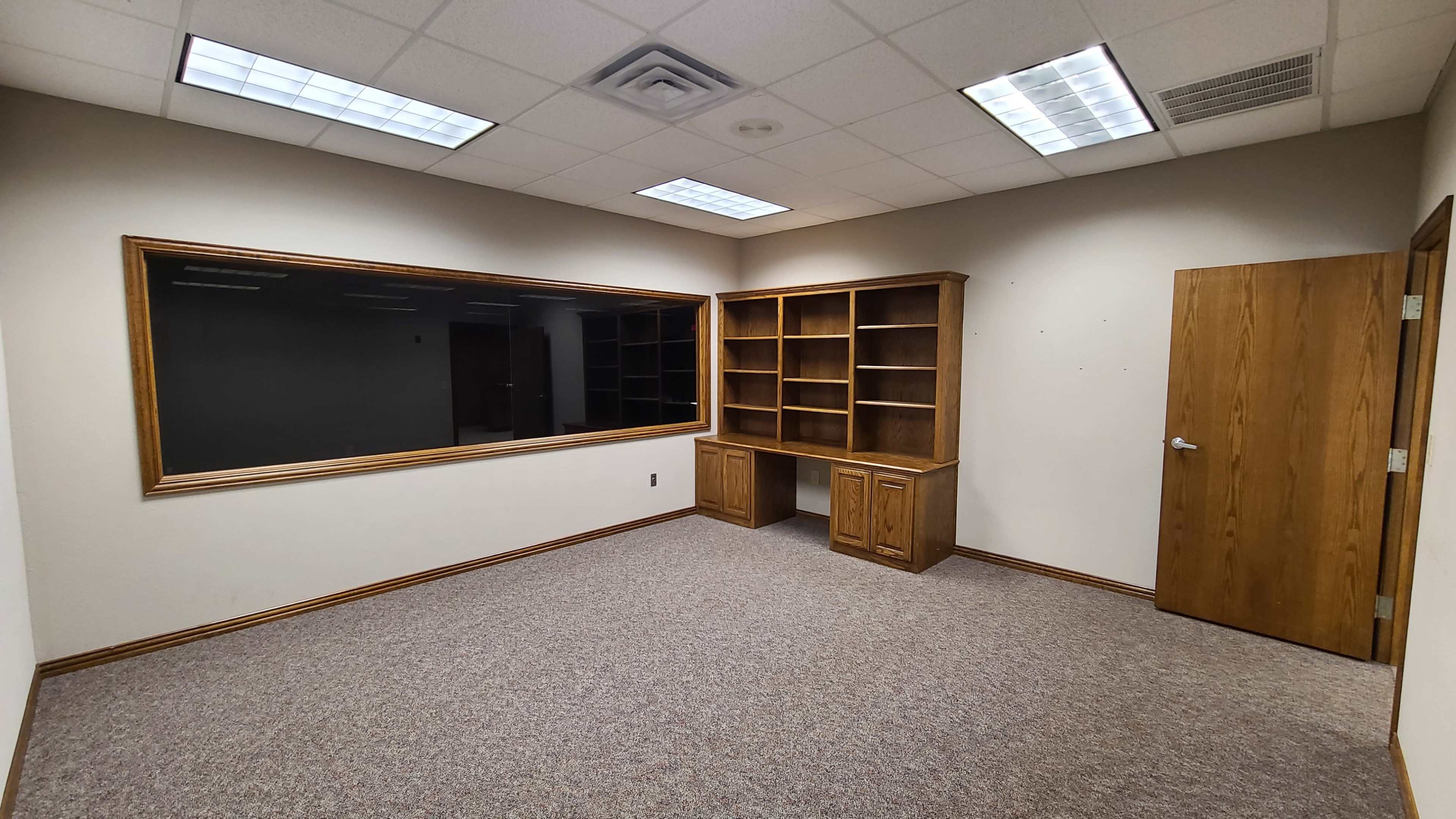 The image shows an empty office space with a built-in wooden bookshelf and a large window mounted on the wall.