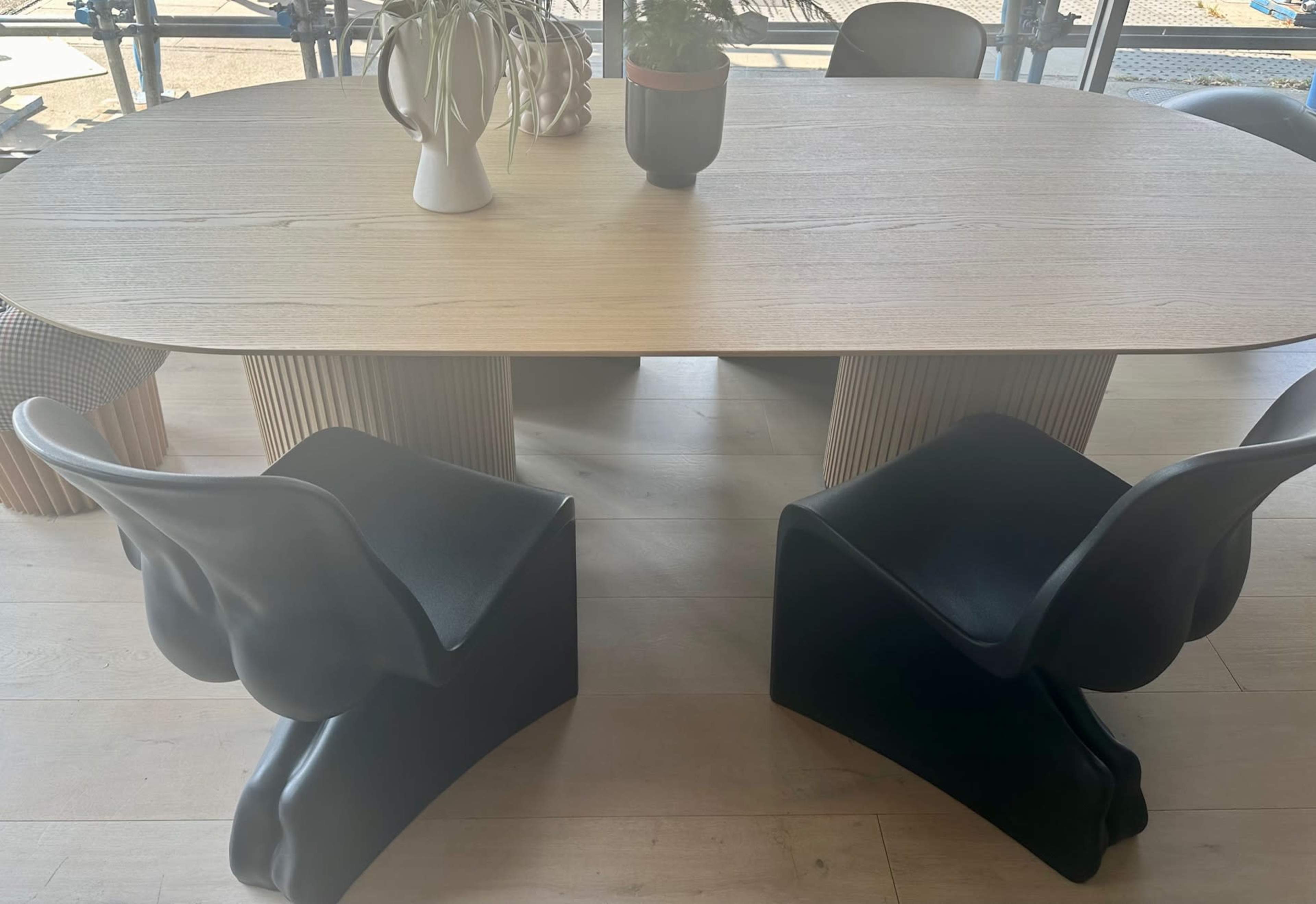 The image shows a modern wooden dining table surrounded by two black sculptural chairs and several plant arrangements.