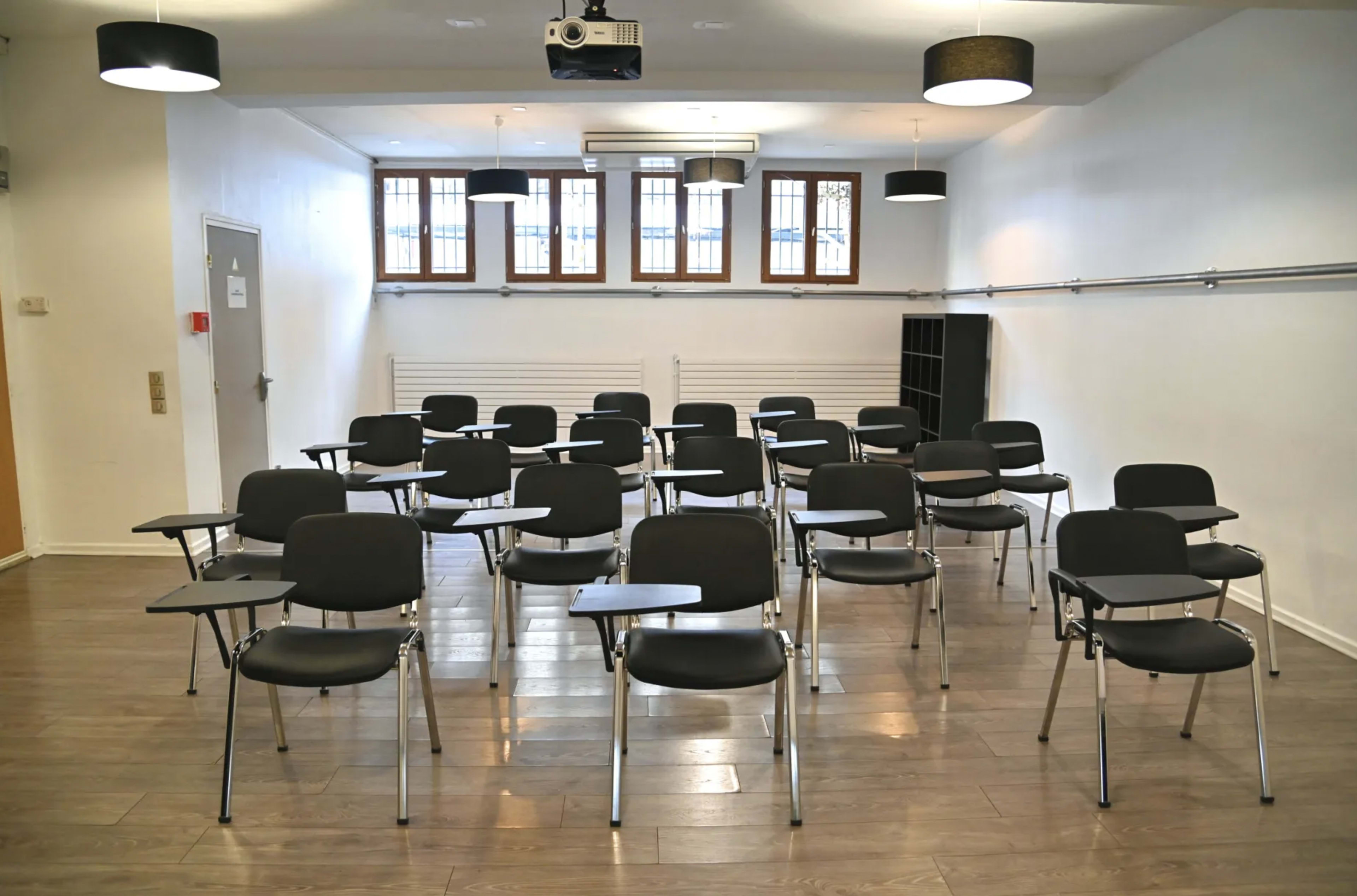 A classroom with rows of black chairs equipped with writing surfaces, arranged neatly on a wooden floor under overhead lighting.
