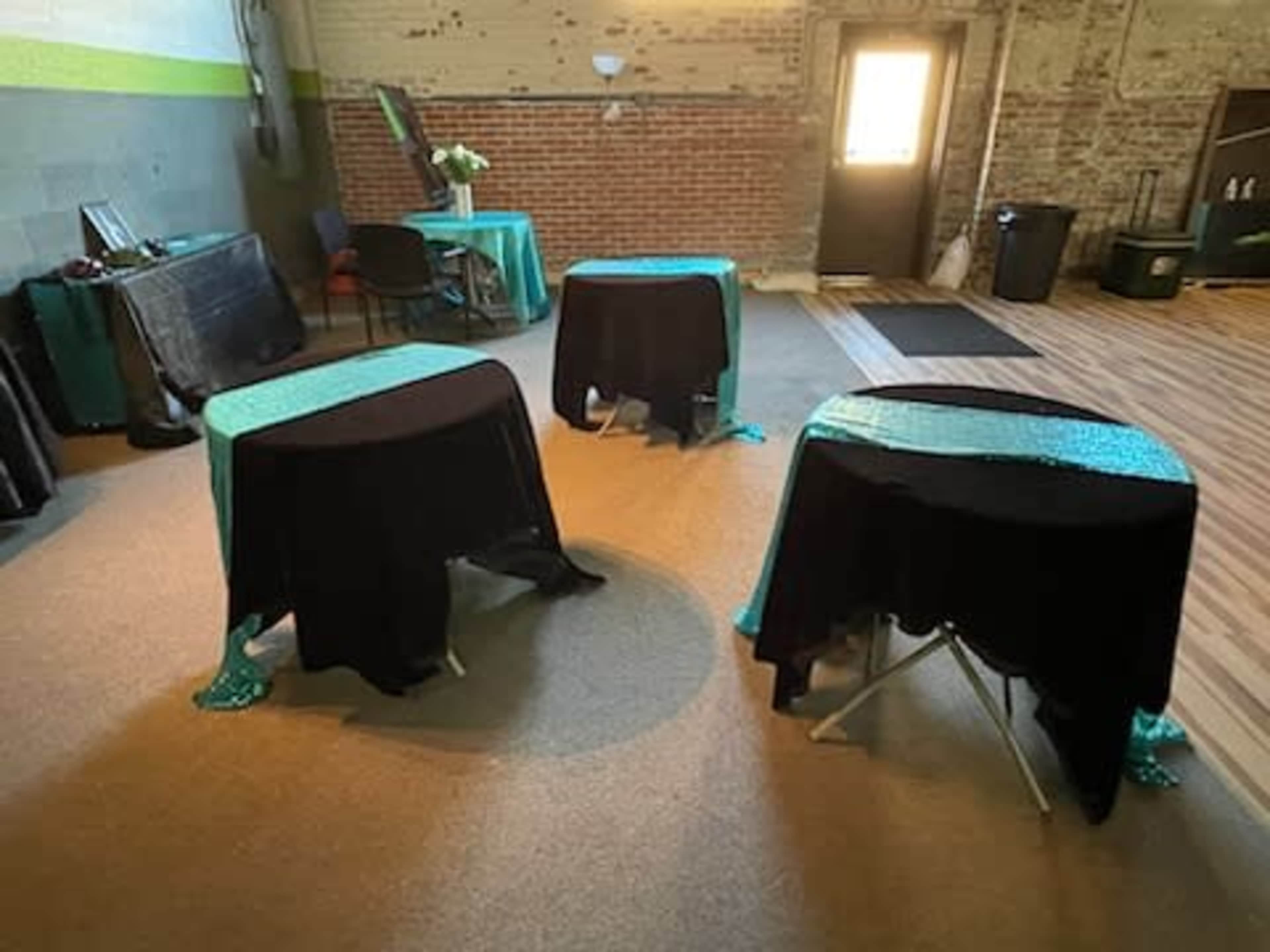 Three tables covered with black and teal tablecloths are arranged in a spacious, brick-walled room with a door leading outside.