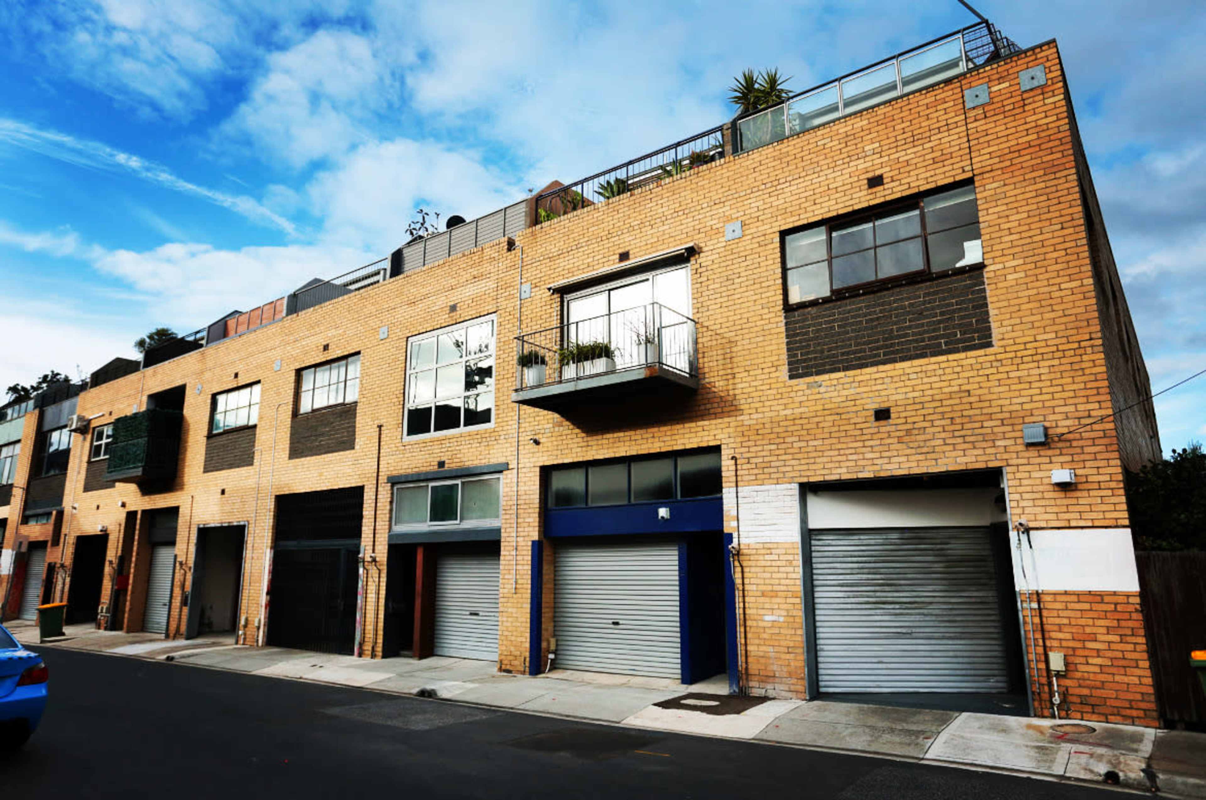 The image shows a brick building with several large windows, a balcony, and multiple garage doors, situated on a street under a partly cloudy sky.