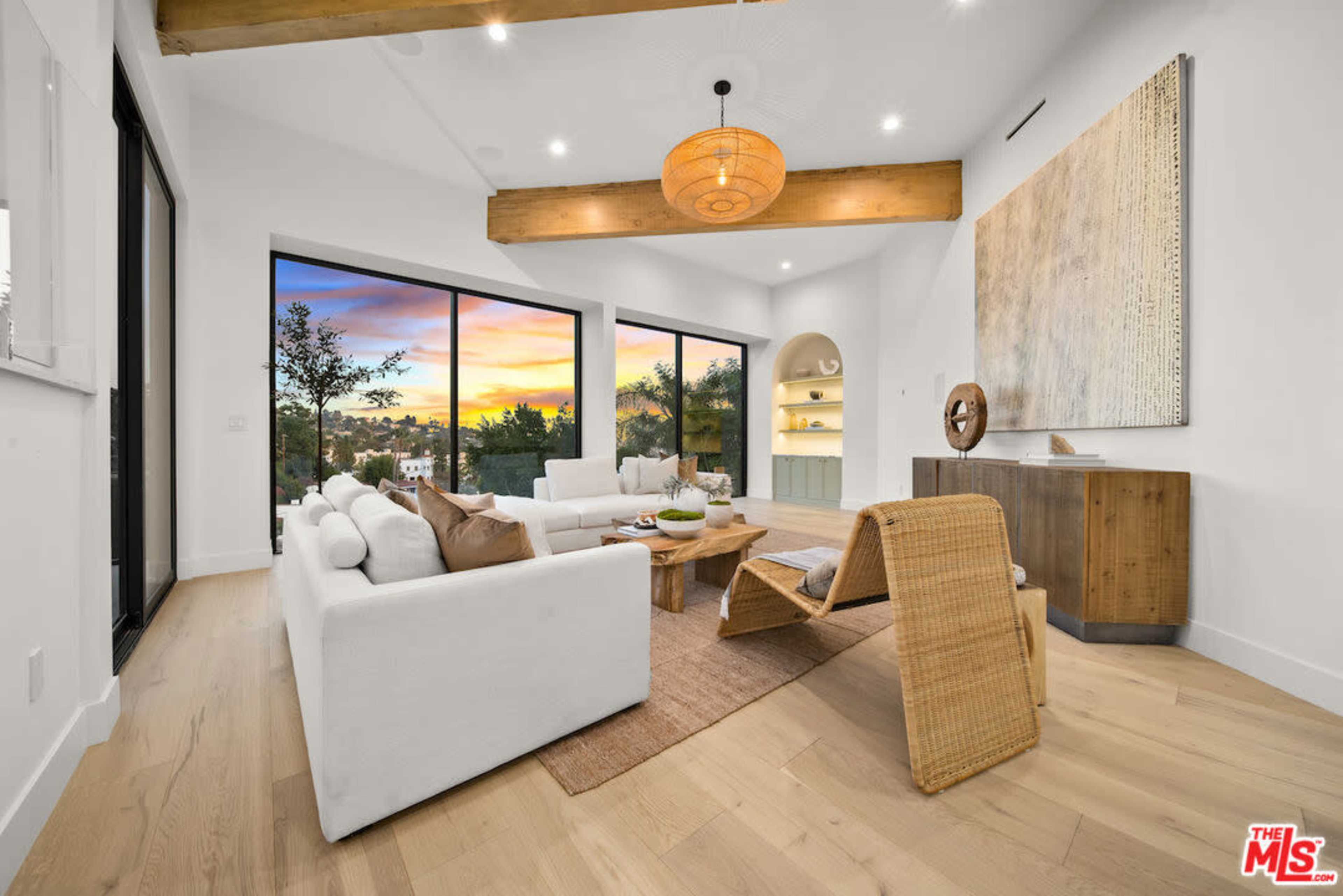 The living room features a light-toned wooden floor, white furniture, large windows showcasing a sunset view, and exposed ceiling beams.
