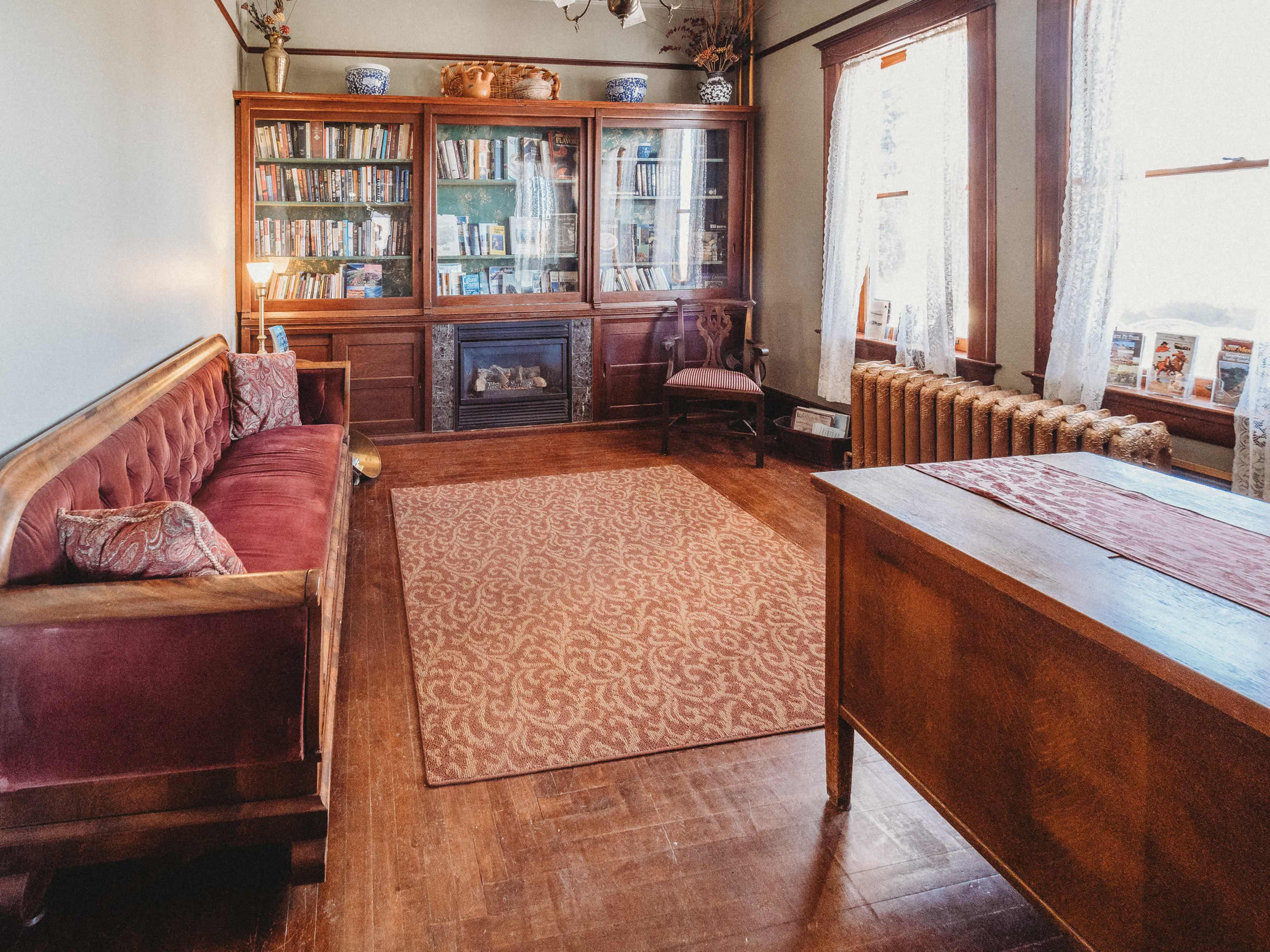 A cozy room with a wooden bookcase, a maroon sofa, an area rug, a wooden table, and a radiator beside large windows.