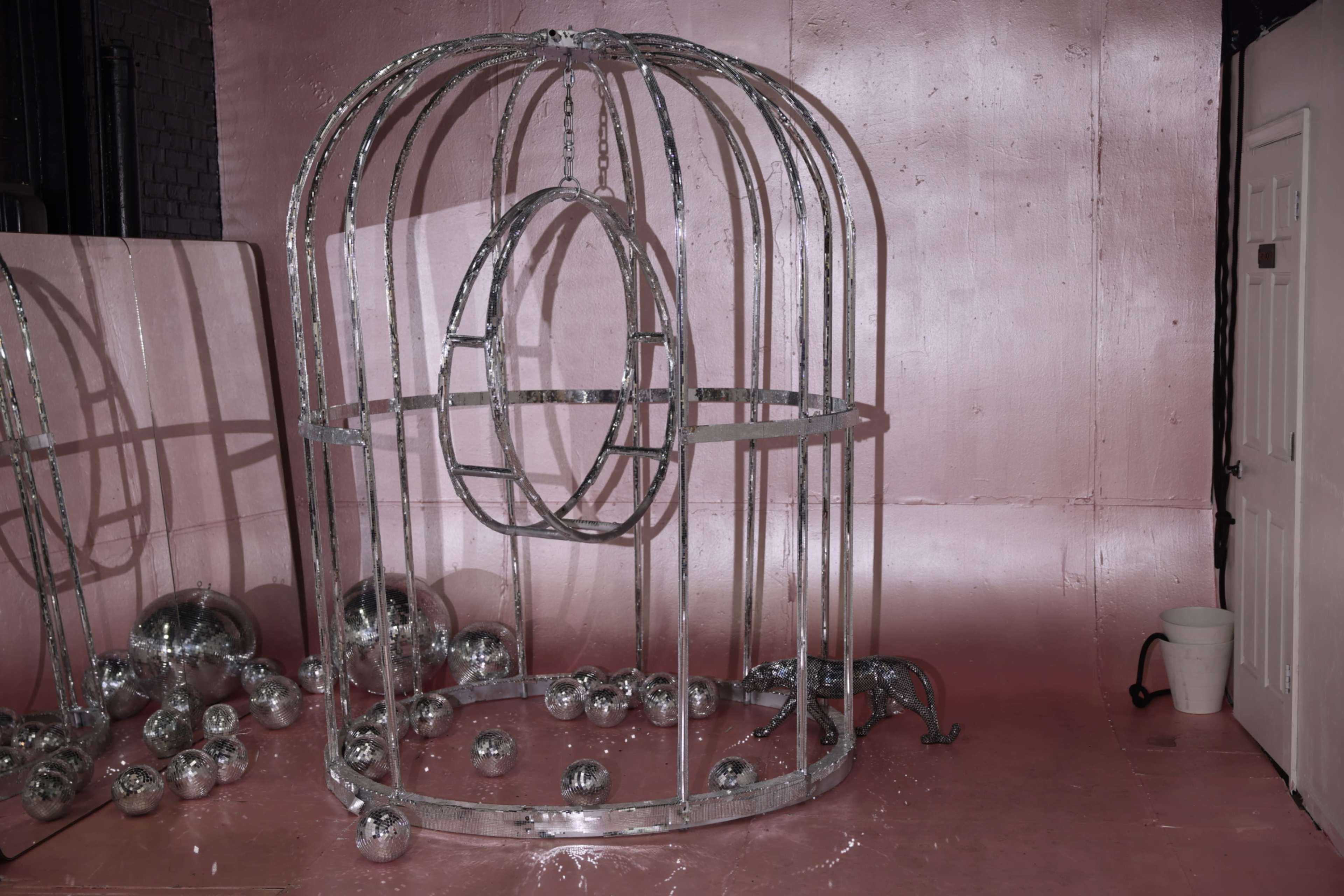 The image depicts a large, metallic cage structure surrounded by scattered reflective balls on a pink floor.