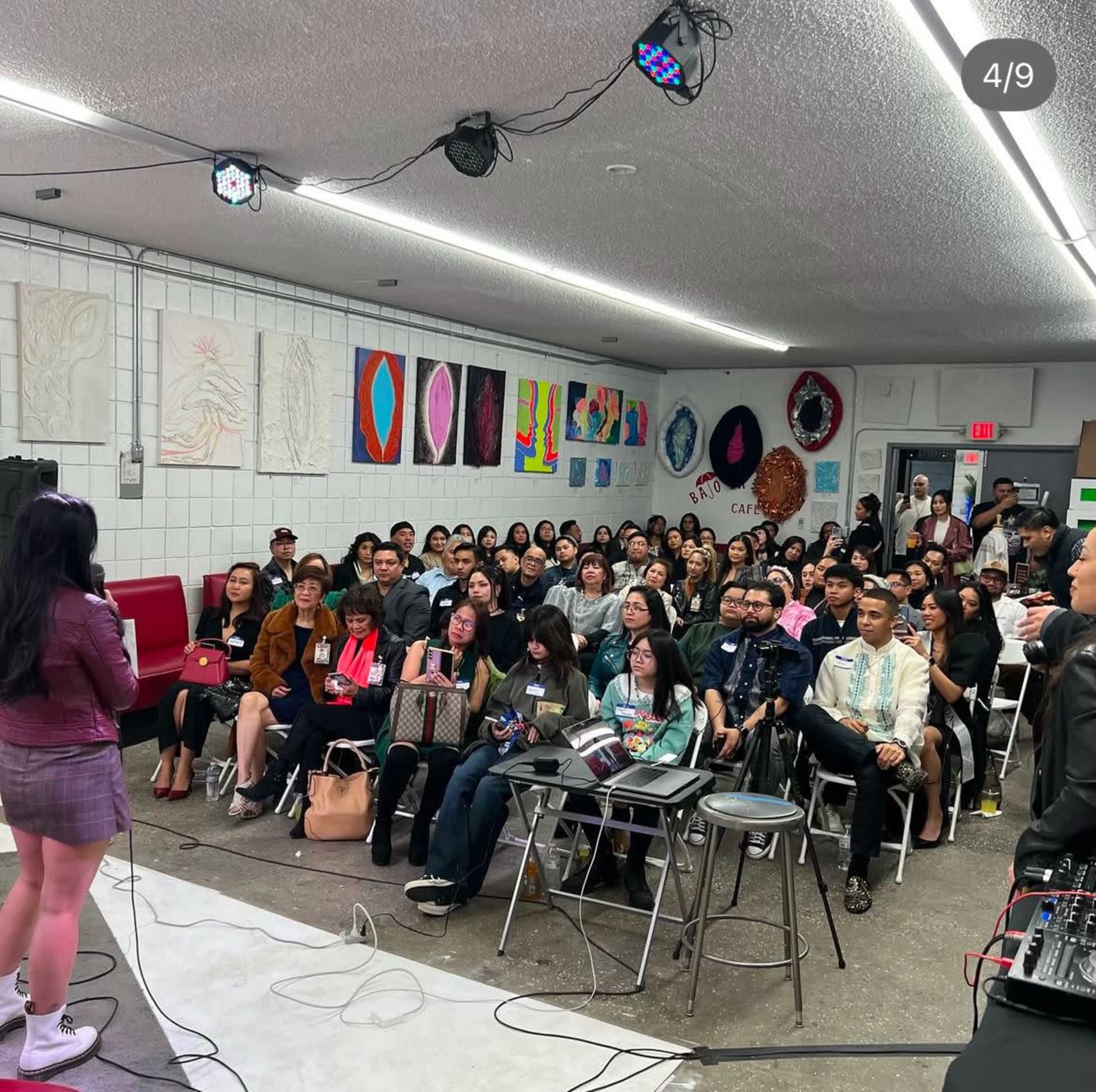 A large audience is seated in a room with colorful artwork on the walls while a speaker addresses them from a small stage.