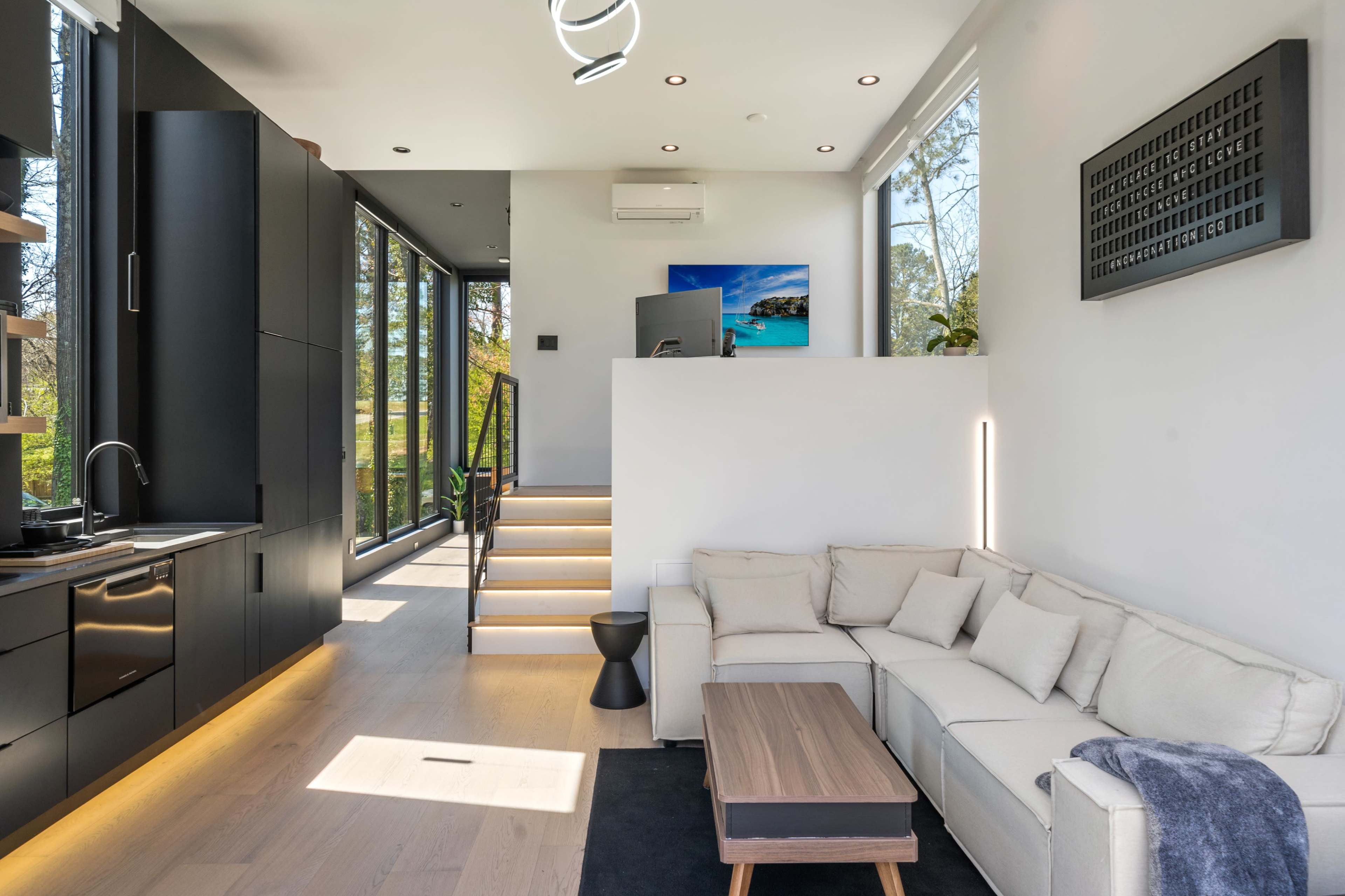 A modern living area with a light-colored sectional sofa, a wooden coffee table, and large windows that provide a view of the outdoors, along with a staircase leading to an upper level.