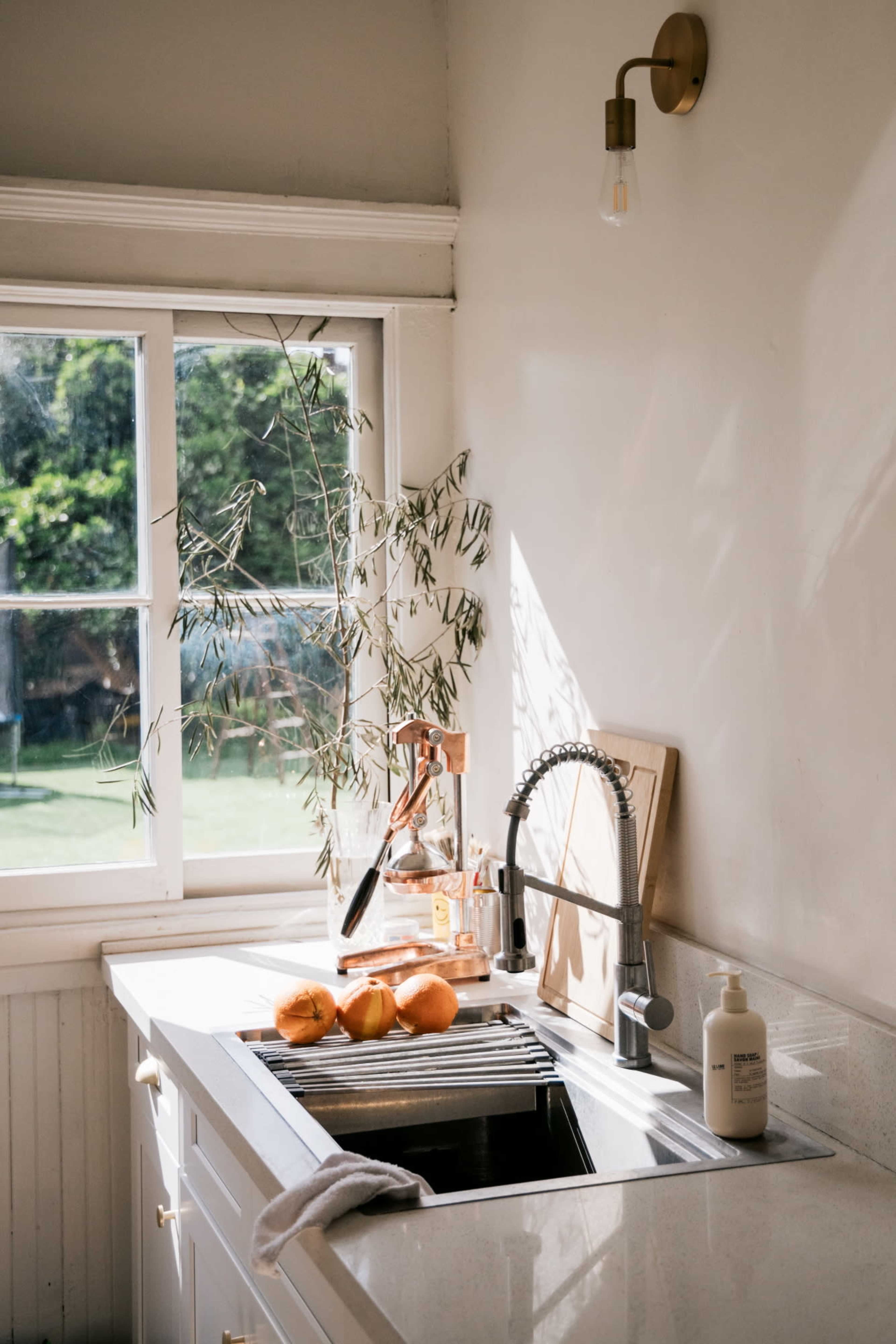 A bright kitchen features a modern sink with oranges and a soap dispenser, illuminated by sunlight coming through a window.