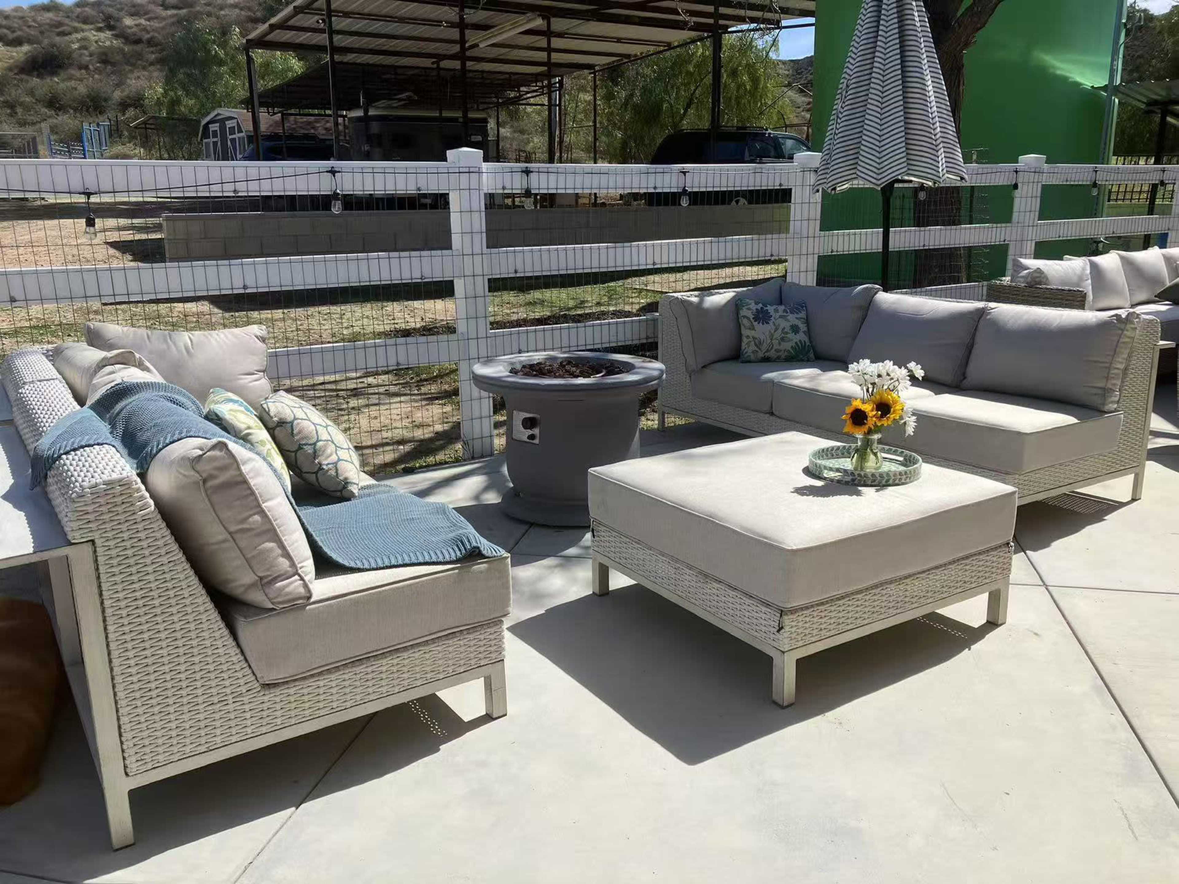 A patio area features a sectional seating arrangement with cushions, a coffee table, and a fire pit, surrounded by a fence and greenery.
