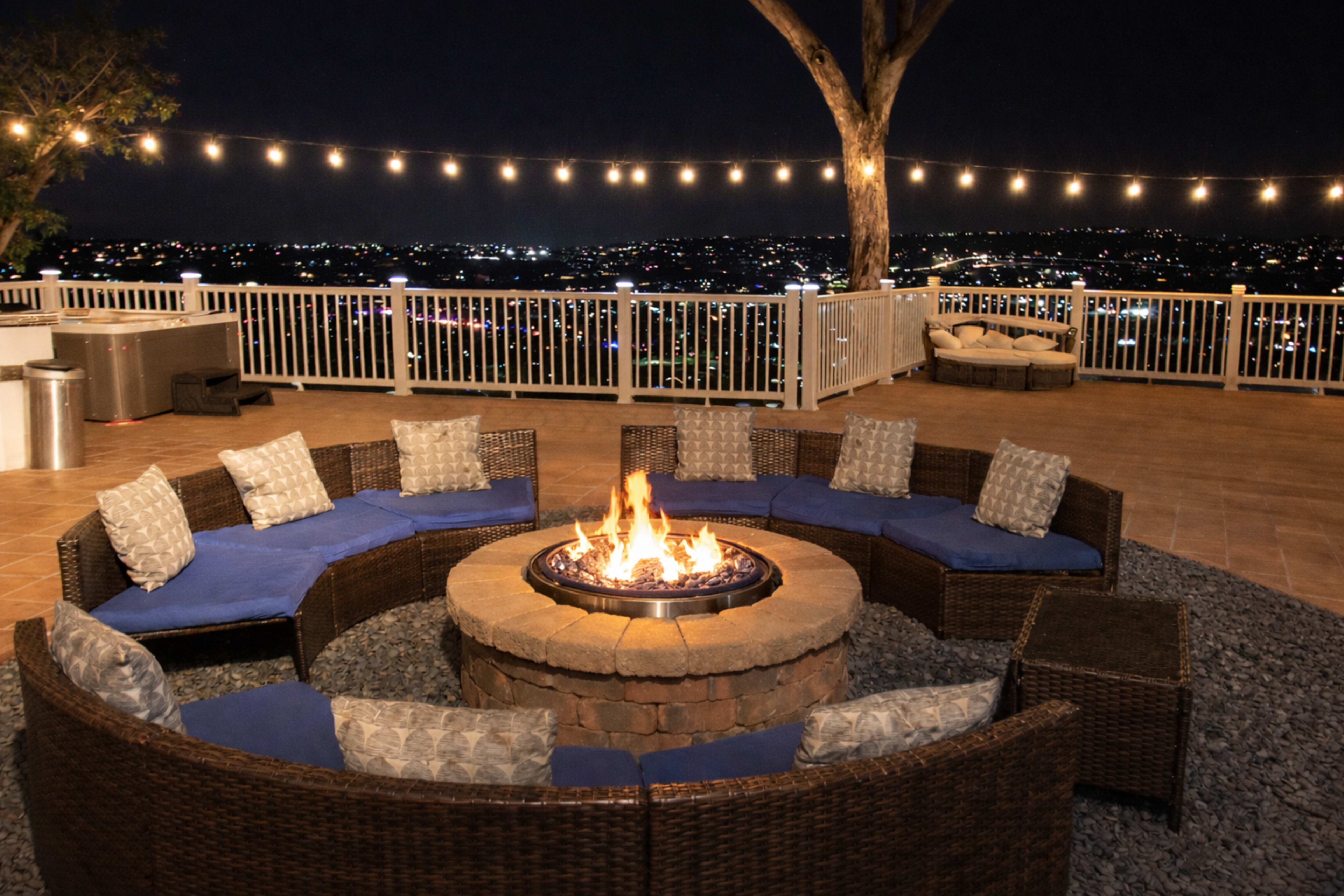 A circular fire pit surrounded by curved wicker seating with blue cushions is illuminated by string lights against a backdrop of city lights at night.