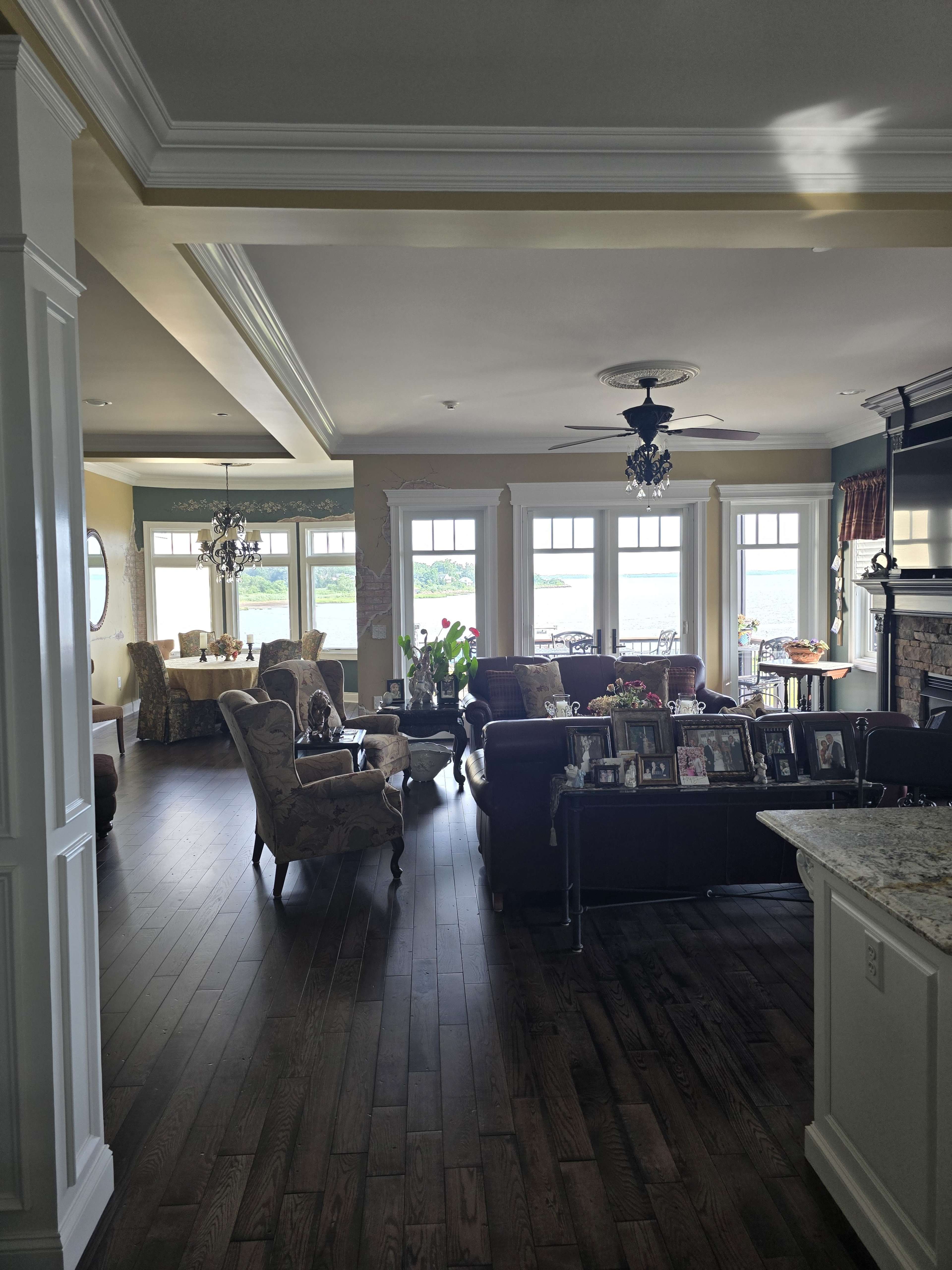 The image shows a spacious living area with hardwood floors, large windows overlooking a water view, and a mix of seating including armchairs and a sofa.