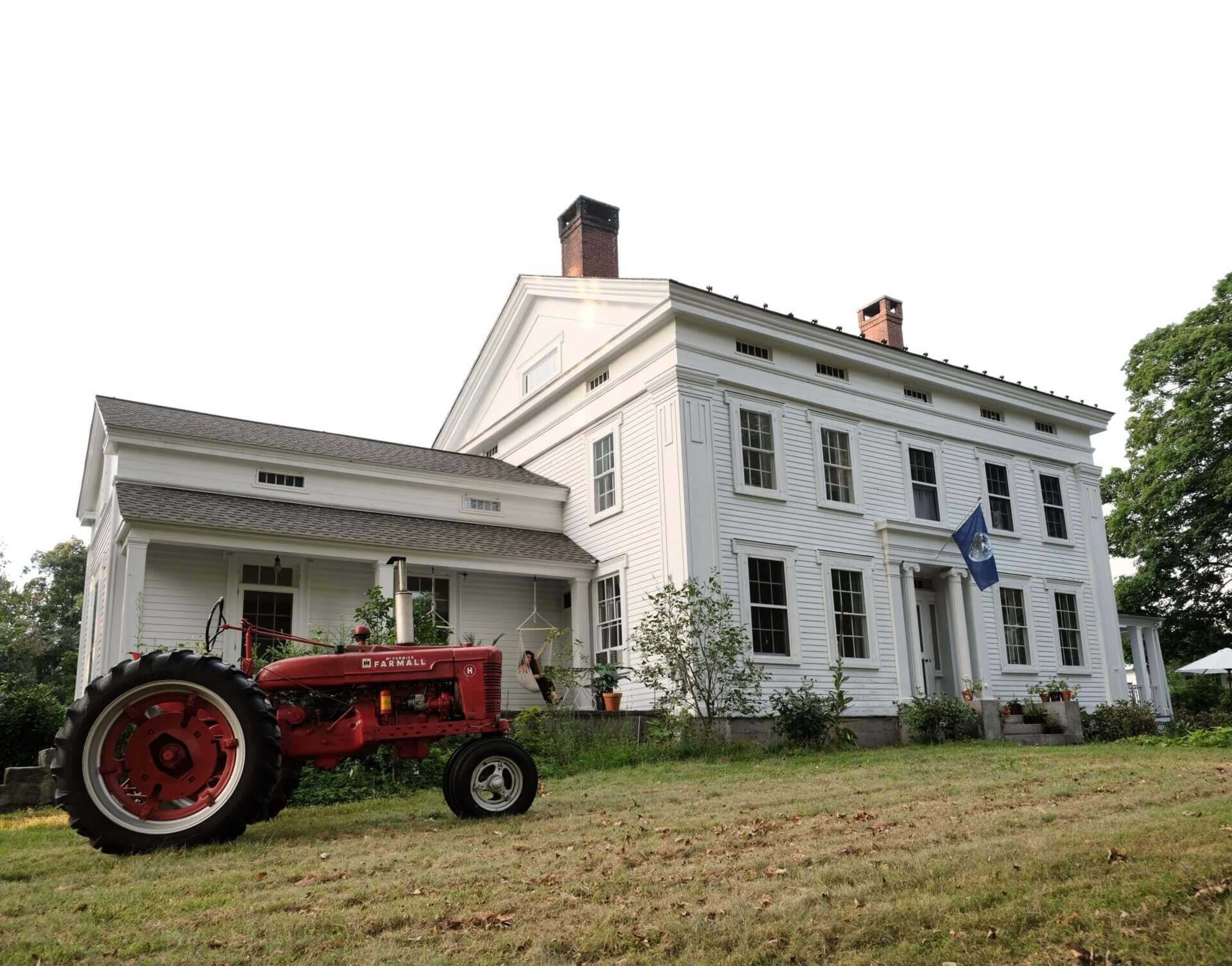 A vintage red tractor rests in front of a large white colonial-style house with a flag displayed.