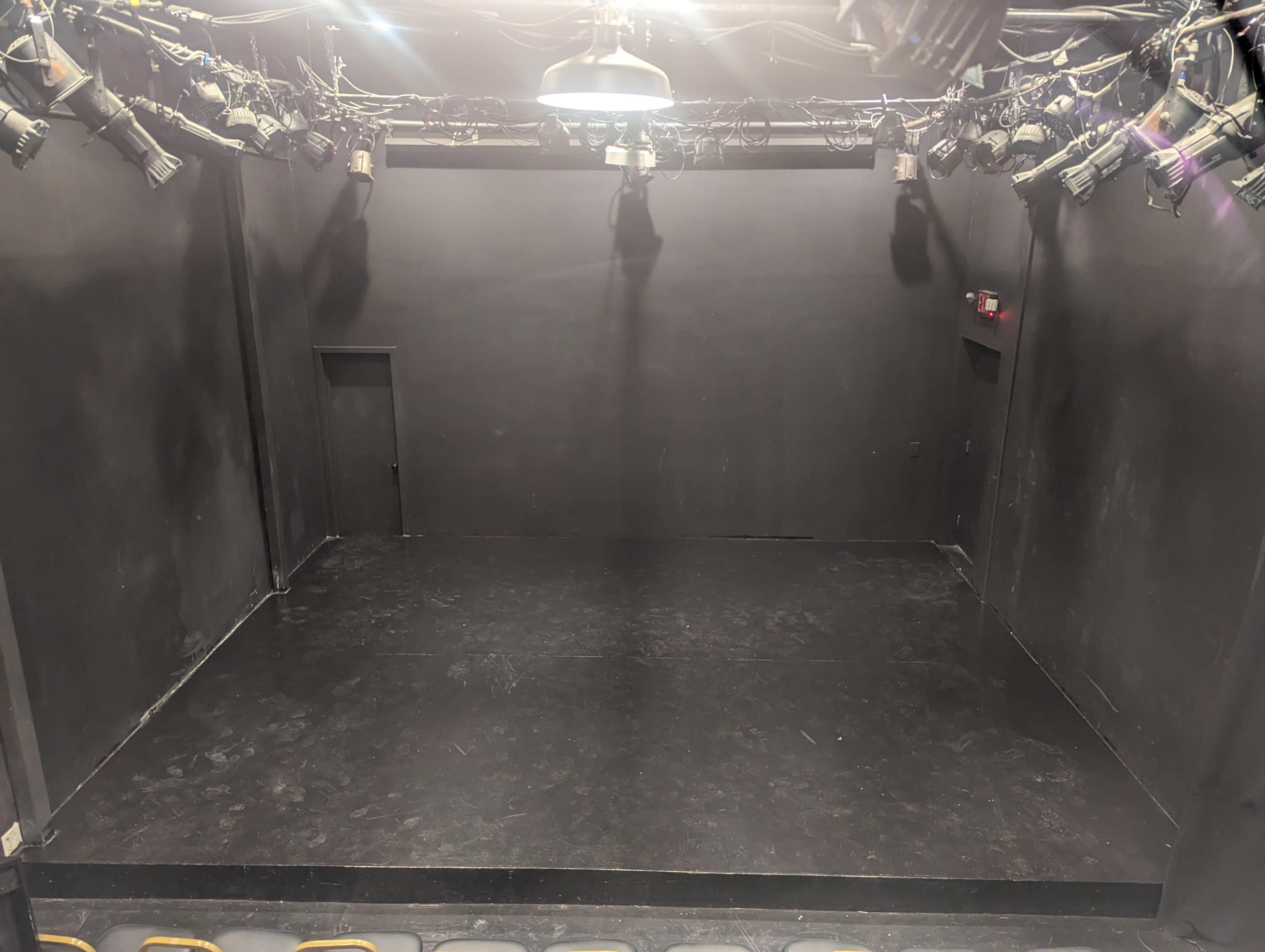 A small, empty black box theater with a few lights hanging from the ceiling and a dark floor.