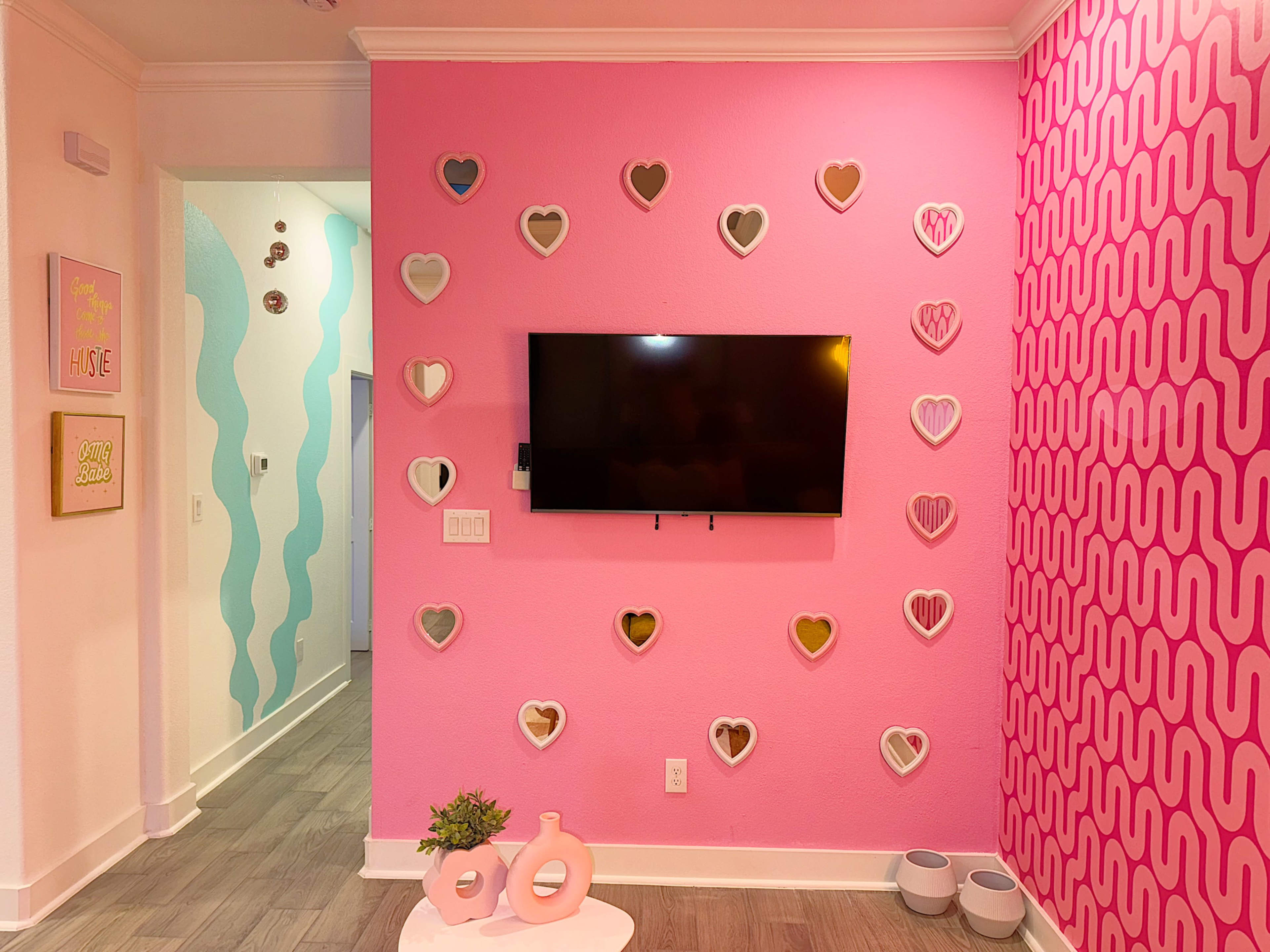 A room with a pink wall featuring heart-shaped frames, a mounted television, and decorative elements on a white coffee table.