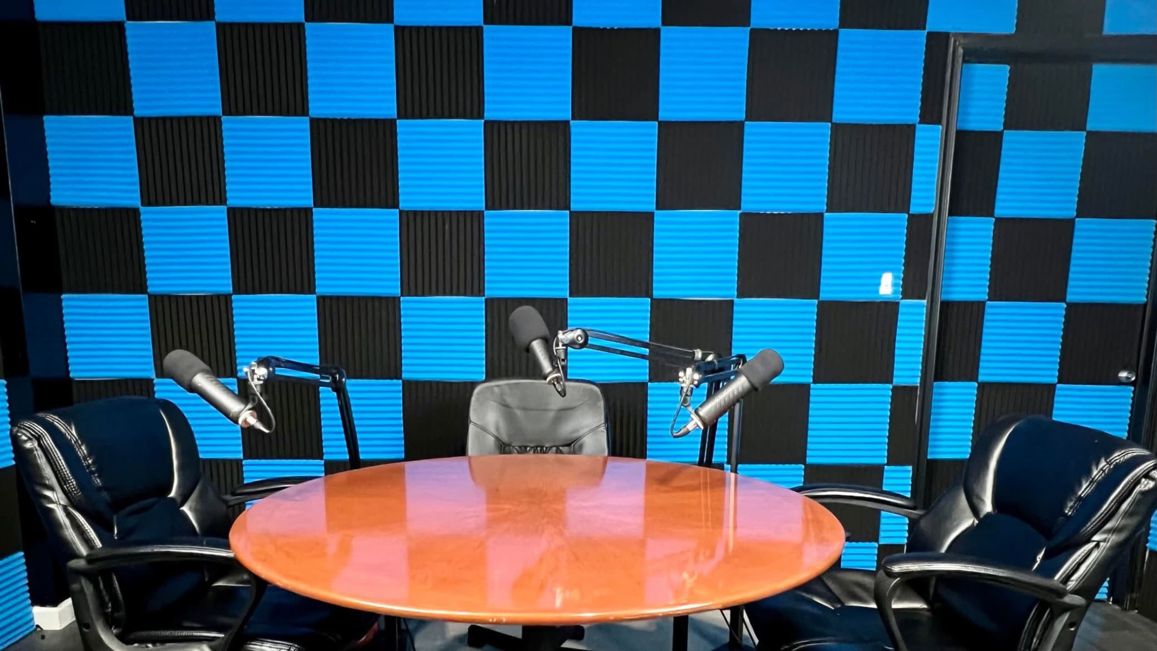 A round wooden table is set in a recording studio with two black chairs and microphones, against a blue and black checkerboard wall.