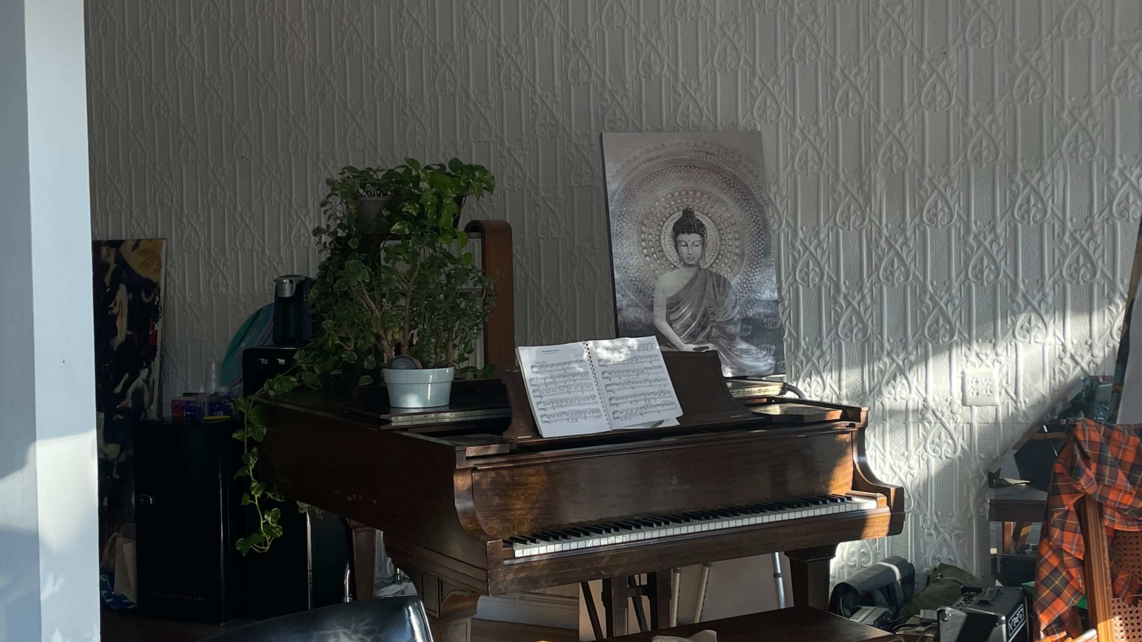 A wooden piano with sheet music sits against a wall adorned with a Buddha artwork and a small potted plant.
