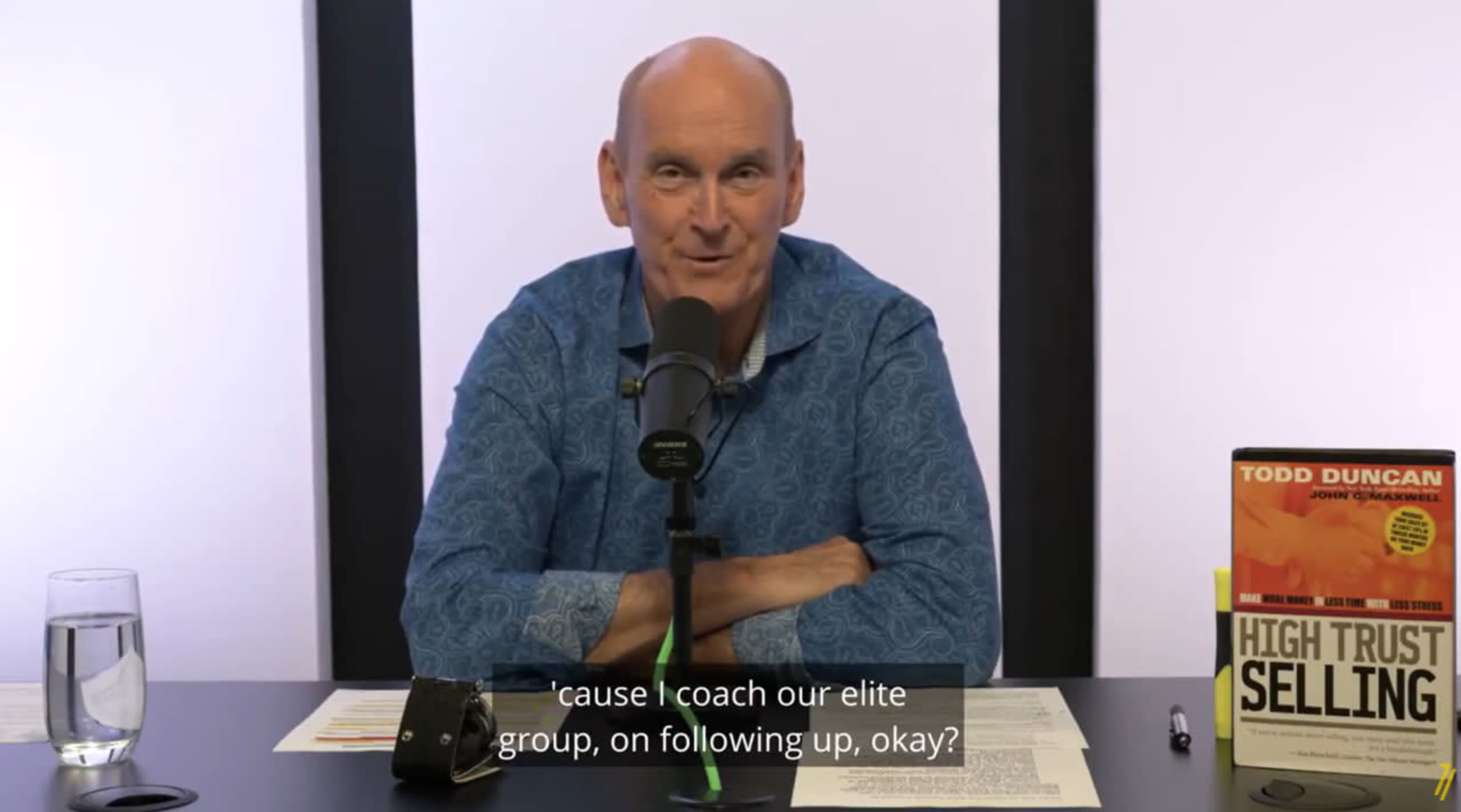 A man sits at a table with a microphone, discussing topics in front of a book titled "High Trust Selling."